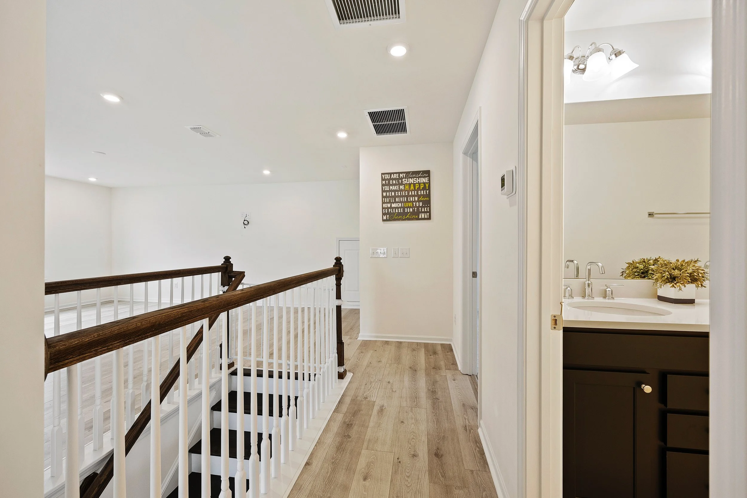 A bright hallway with light wood flooring, white walls, and a staircase with a dark wood handrail and white balusters. To the right, part of a bathroom with a dark vanity, white countertop, and a plant. A decorative sign with a motivational message h
