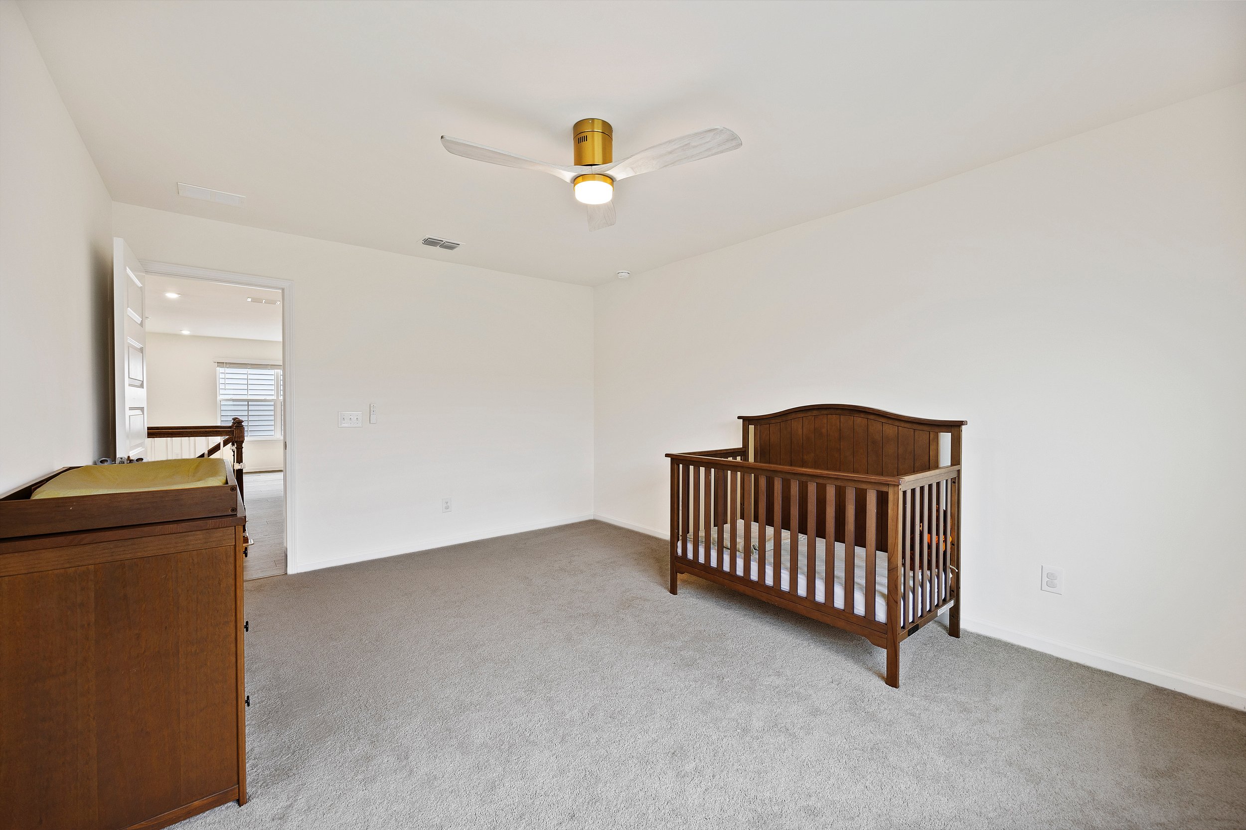 Empty nursery room with white walls, beige carpet, a wooden crib, a wooden changing table, and a ceiling fan.
