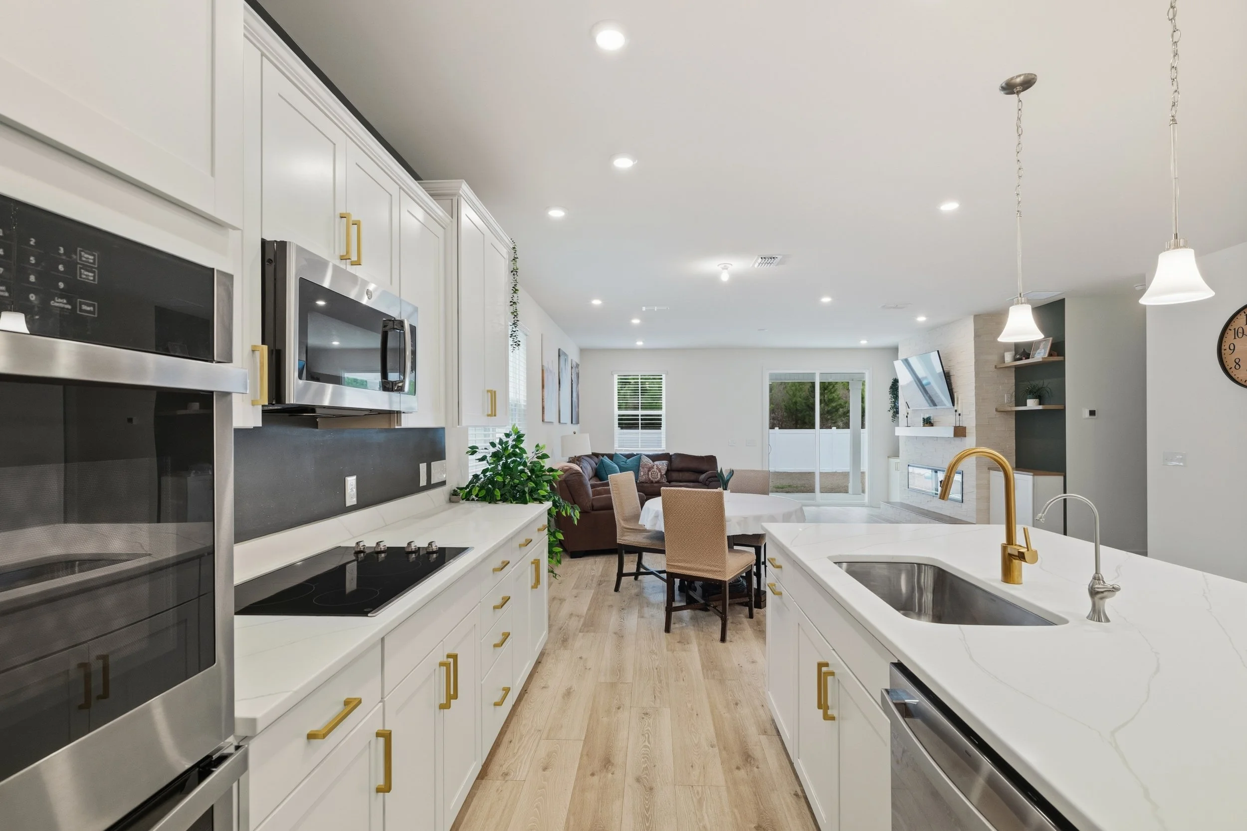 Open-concept kitchen and living room with white cabinets, granite countertops, stainless steel appliances, a dining table, a brown sectional sofa, and a fireplace with a mounted TV.