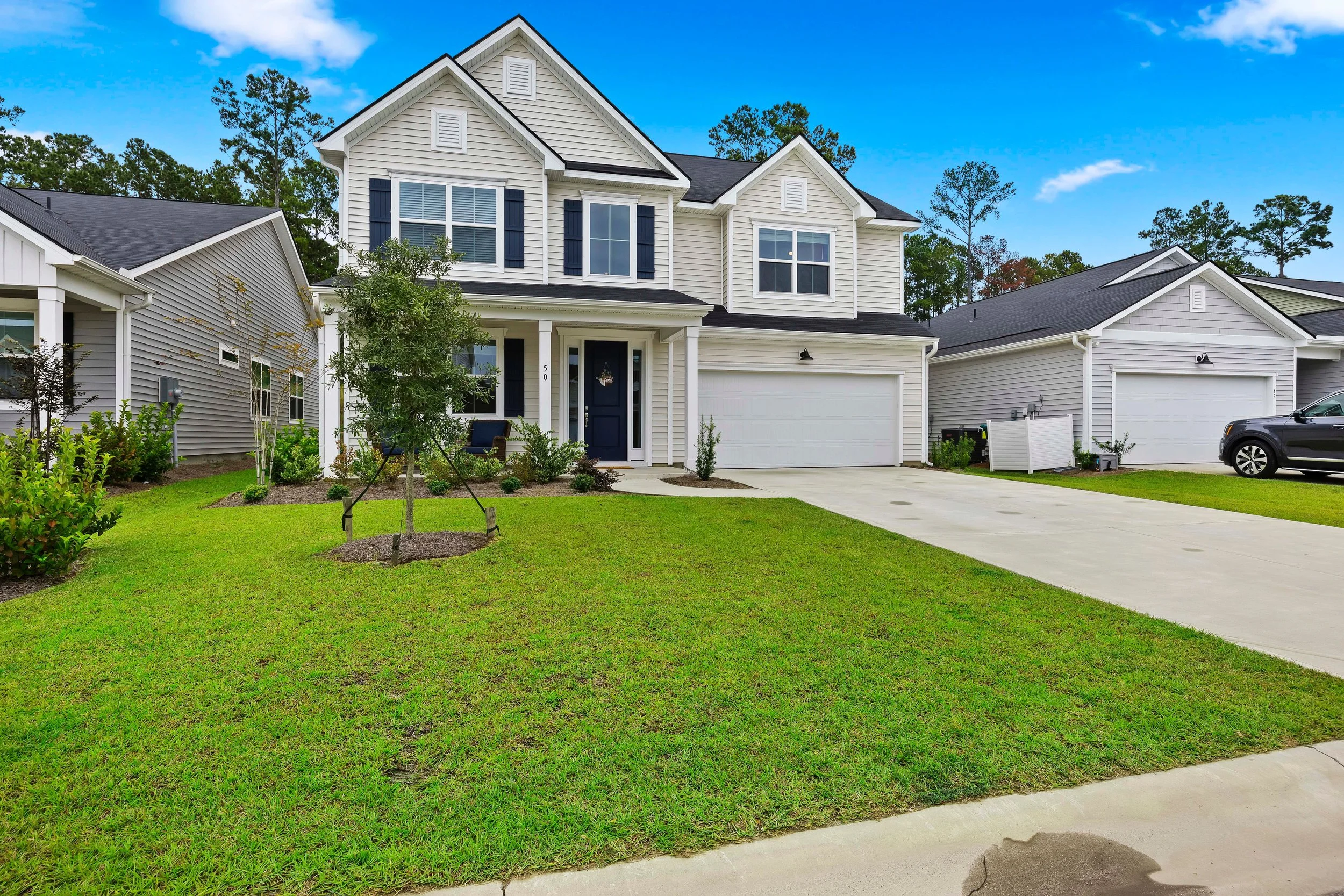 A two-story white house with black shutters, a garage, and a front lawn with small trees and plants.