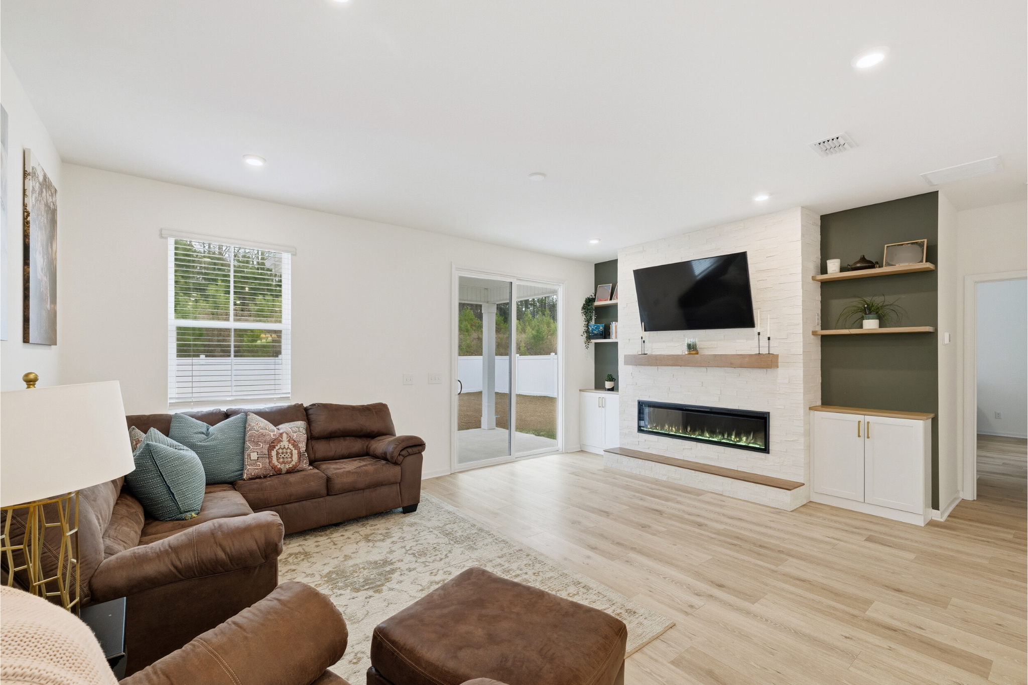 Living room with brown leather sofas, a rug, a window with white blinds, a sliding glass door, wall-mounted TV, a modern fireplace, and open shelving on a white brick accent wall.
