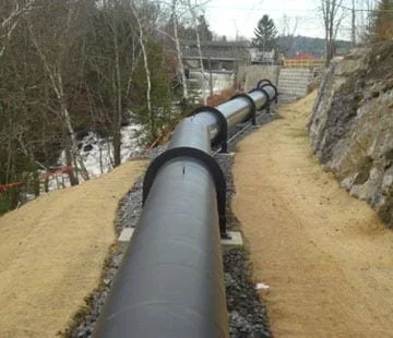 Large black pipe installed along a dirt pathway next to a rocky hillside, with a bridge and trees in the background.