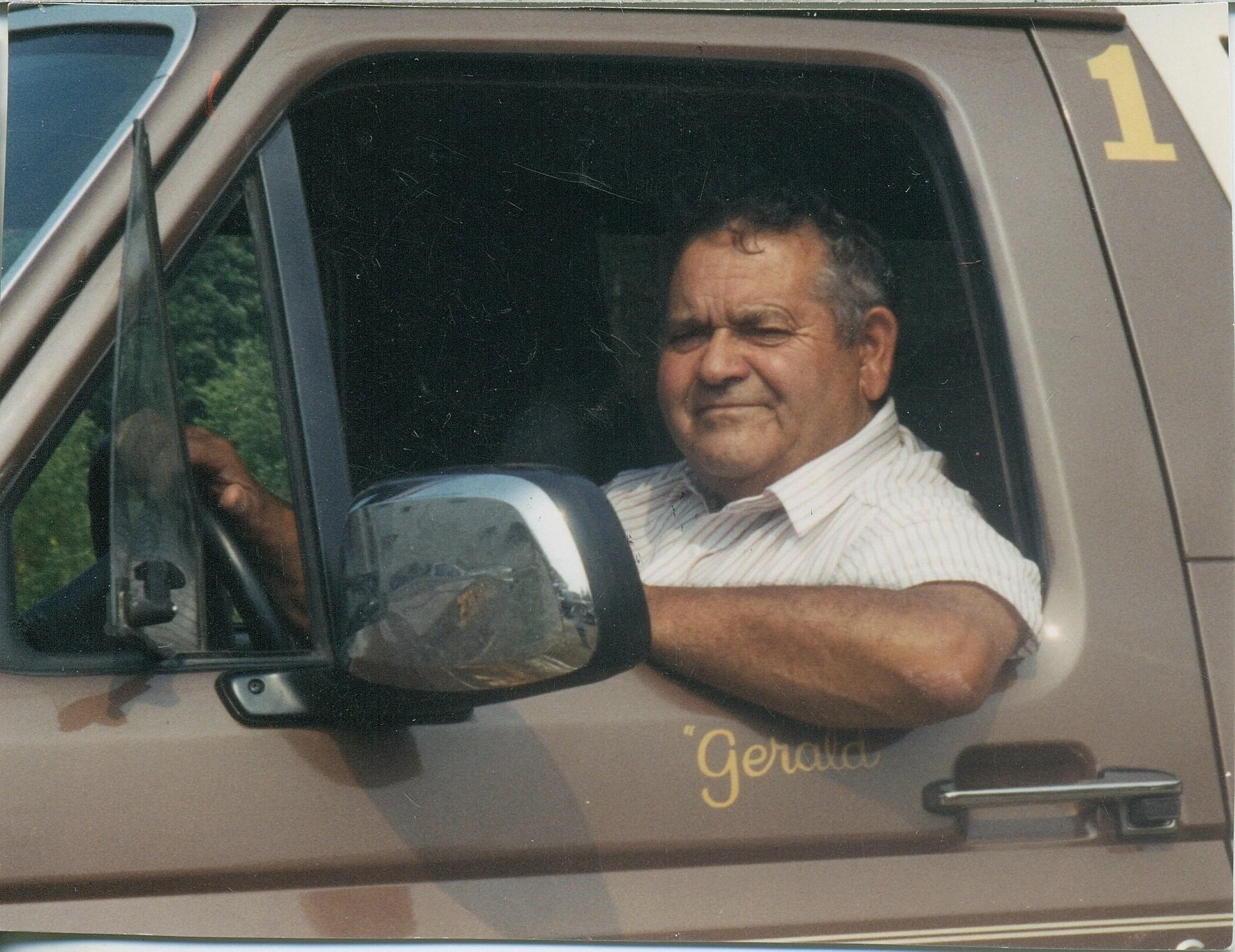 A middle-aged man with short, dark hair and a tan complexion sitting in the driver's seat of a vehicle with his arm resting on the window frame, looking at the camera. The vehicle appears to be a beige or light brown van or truck, and has the name 'Gerald' written near the door.