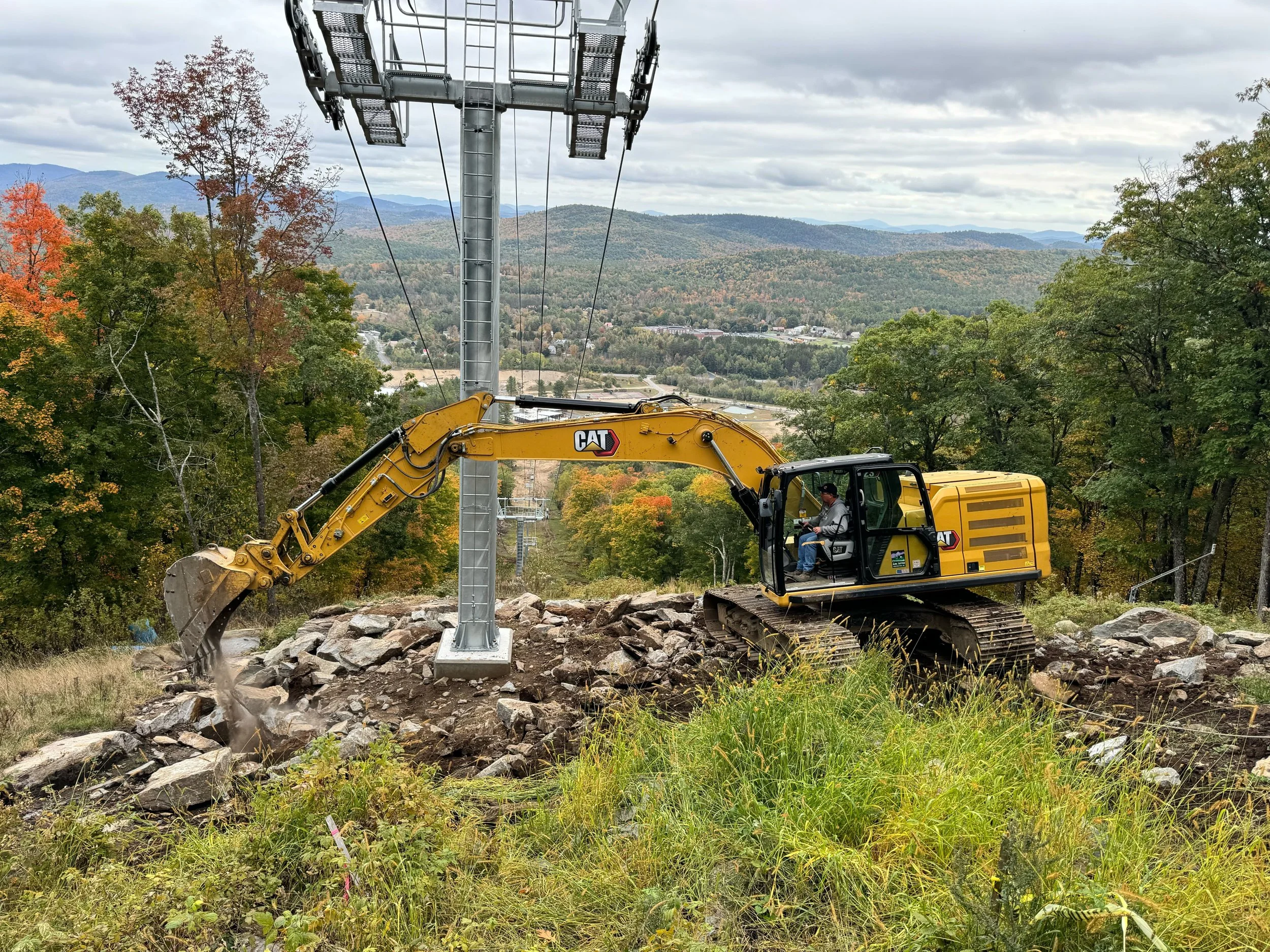 A yellow CAT excavator is demolishing a rocky surface on a hillside, with trees and mountains in the background under cloudy skies.