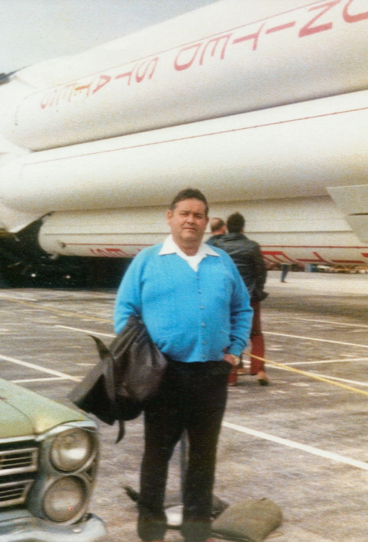A man in a blue sweater and black pants standing in a parking lot near a vintage car, with a large boat or ship in the background, and other people nearby.