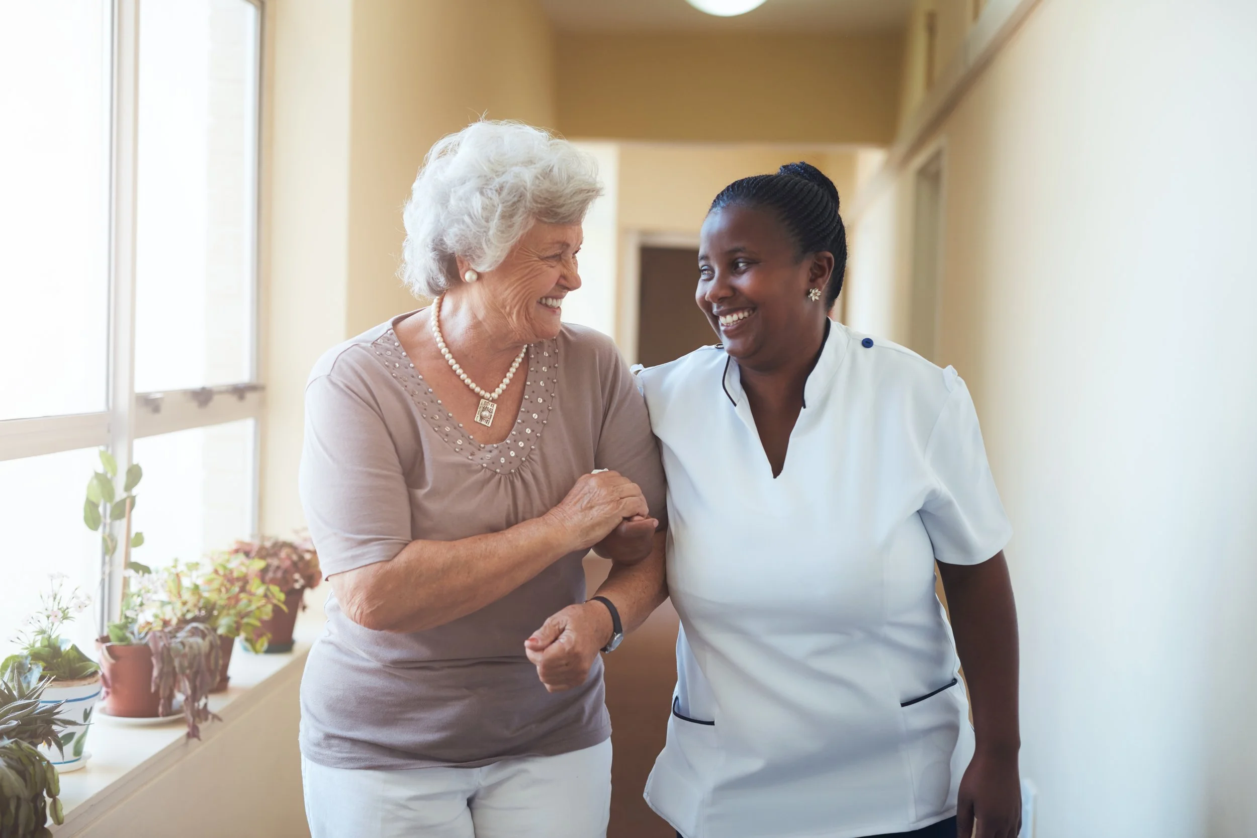 An elderly woman and a female healthcare professional walking together in a hallway, smiling and engaging in friendly conversation.