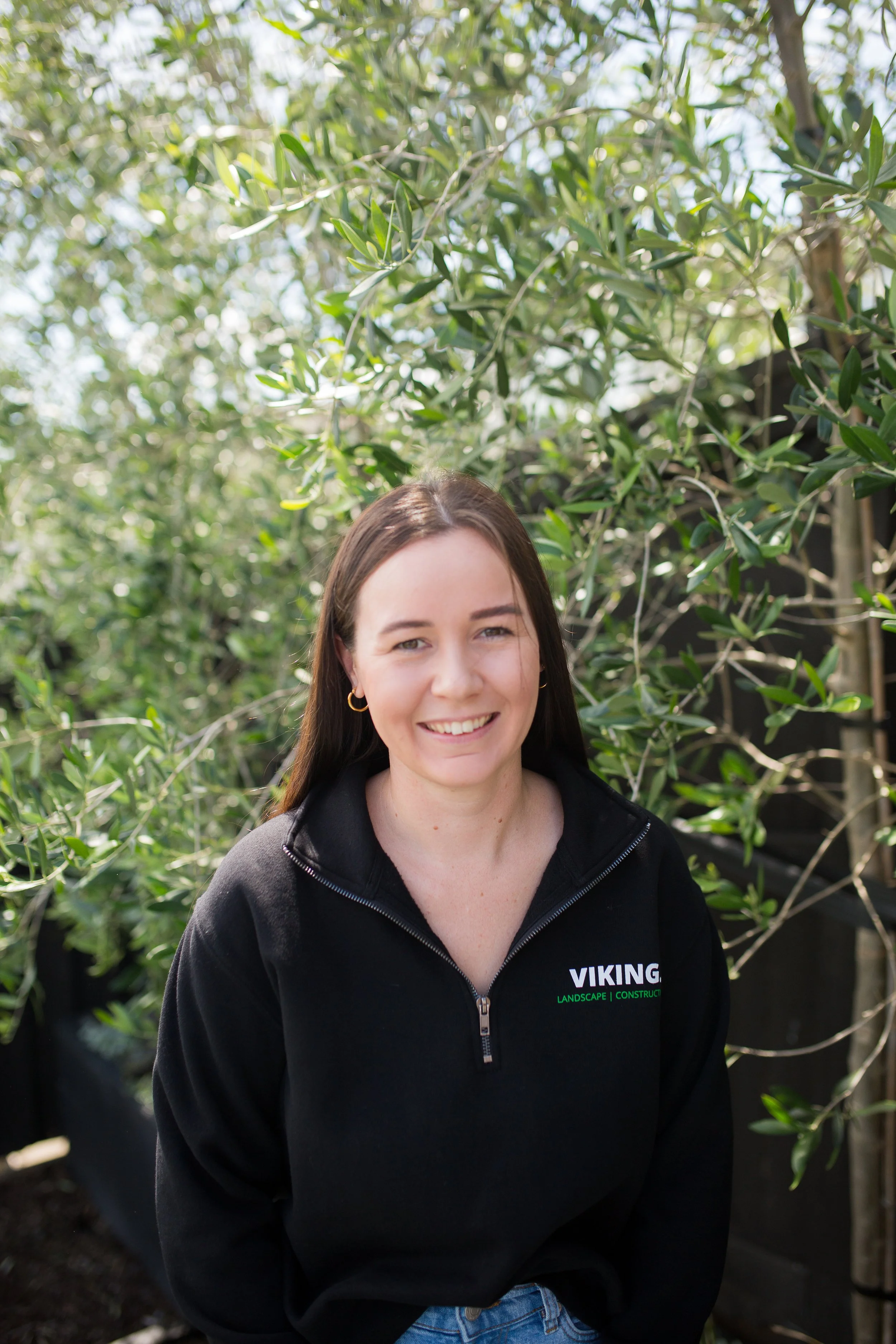 A woman with long brown hair smiling outdoors, wearing a black Viking Landscape and Construction hoodie, standing in front of green leafy plants.