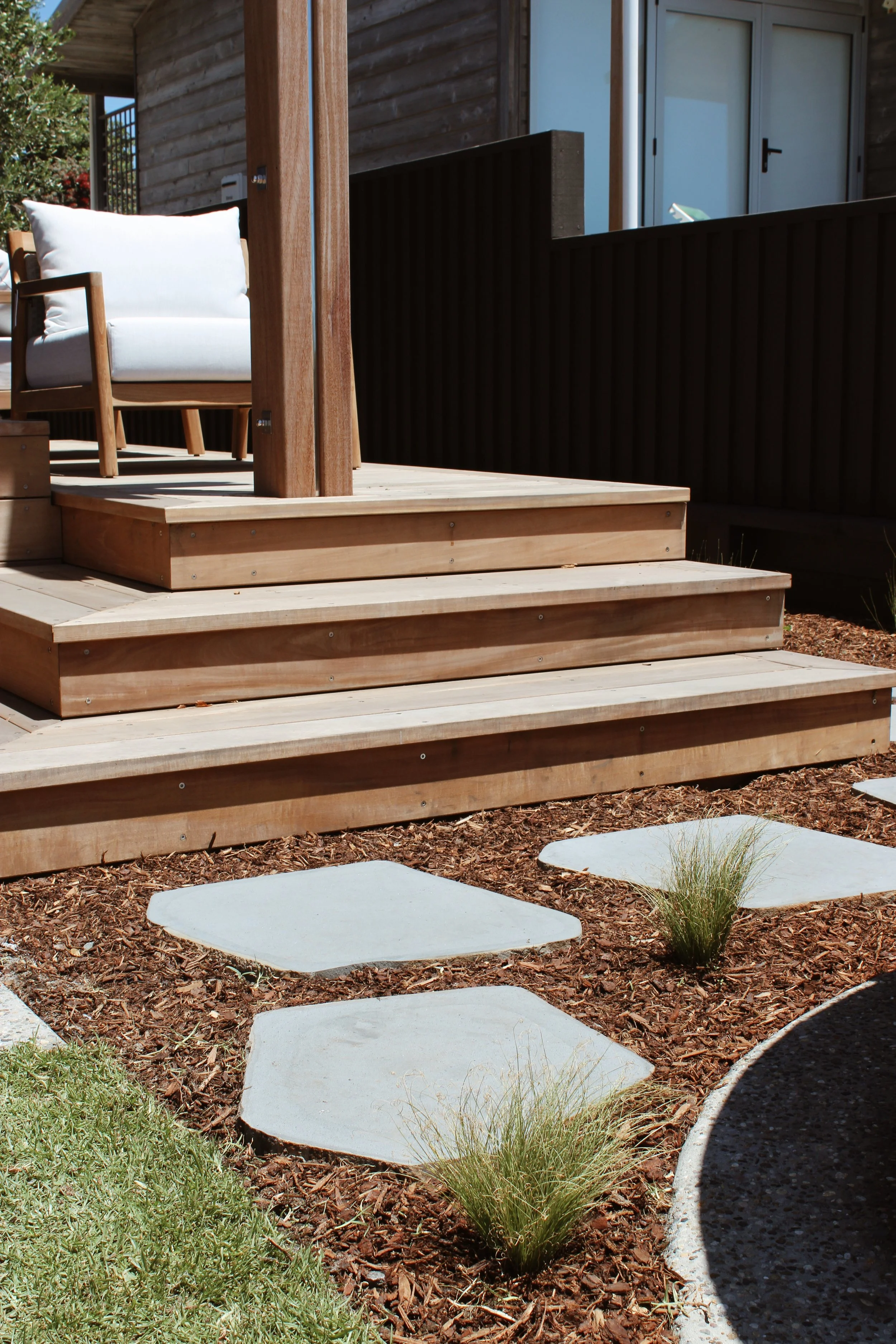 Wooden deck with a white cushioned chair, stepping stones in the garden, and a modern house in the background.