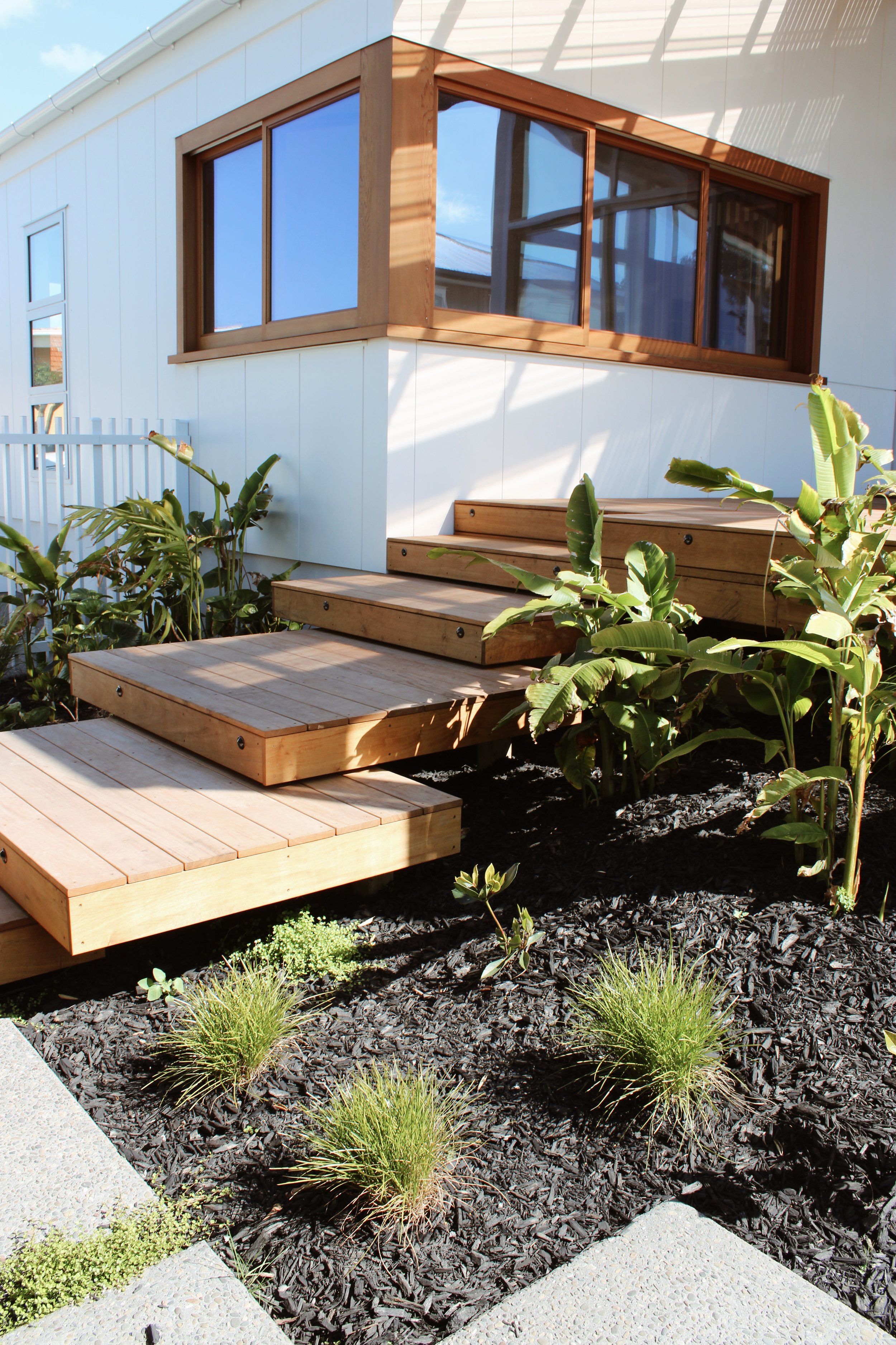 Modern house exterior with large windows, wooden steps, and a landscaped garden with small plants and dark mulch.