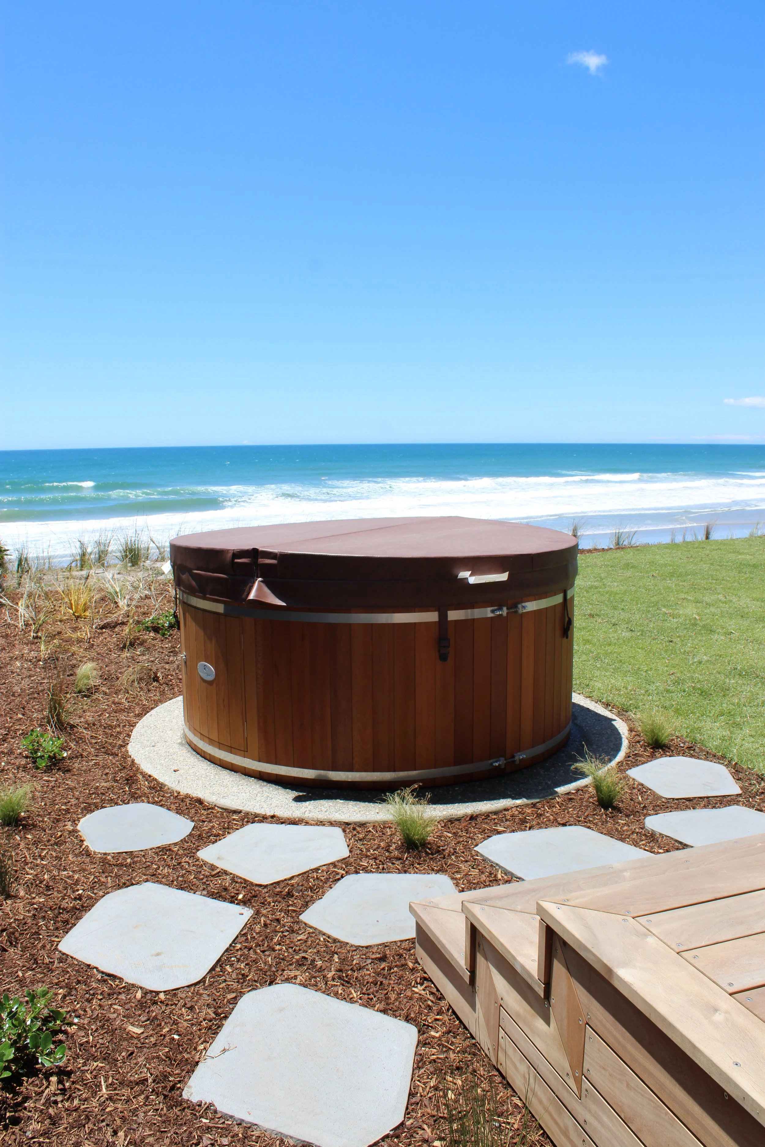 A round outdoor hot tub on a patio overlooking the ocean with a clear blue sky. There are stepping stones in the garden surrounding the tub and wooden effort benches nearby.