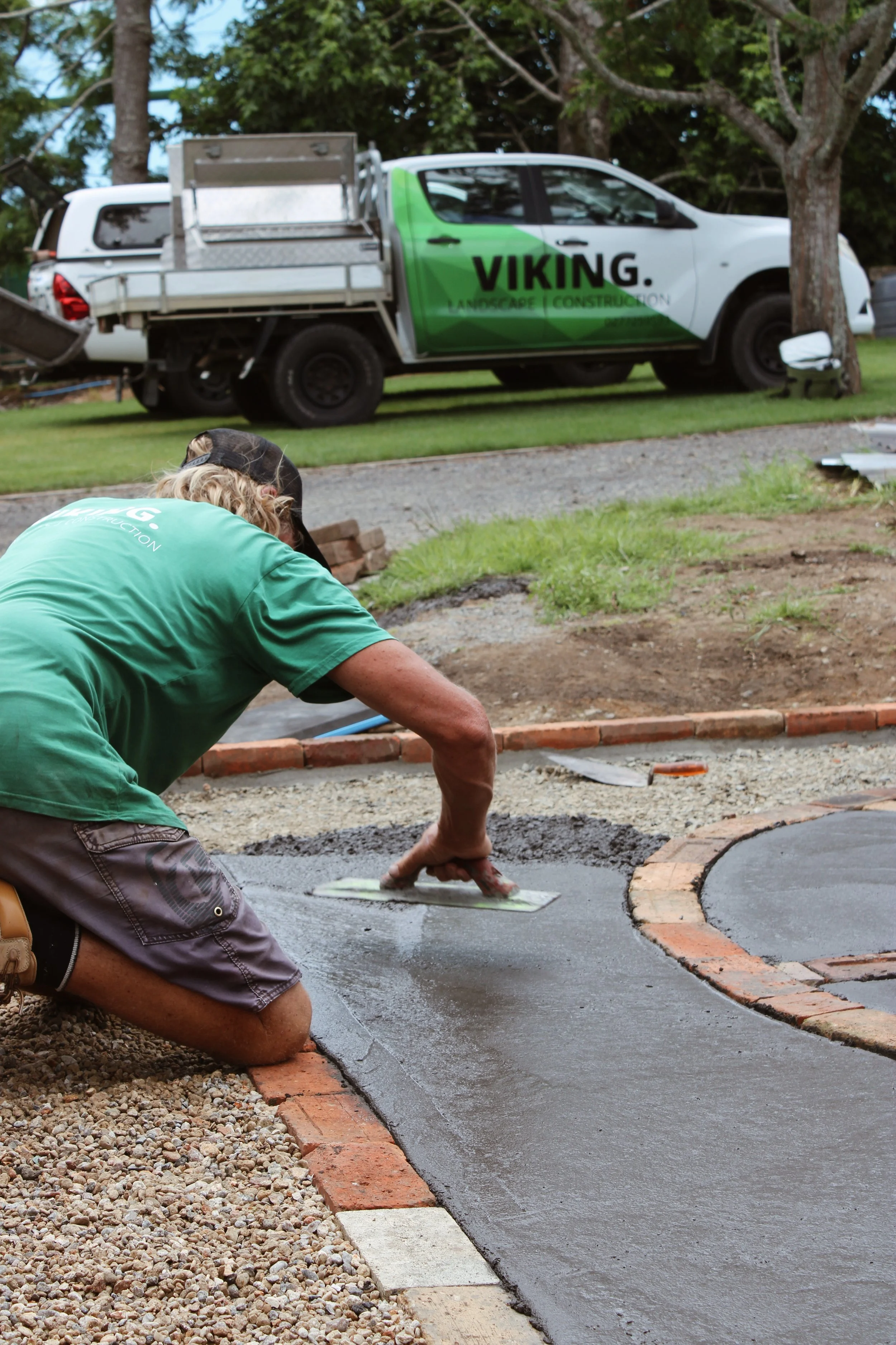 A person kneeling on a gravel surface, smoothing fresh concrete on a paved pathway, with a blue trowel in hand, near a partially completed brick border, while a white pickup truck with green and black branding for Viking Landscape Construction is parked nearby under trees in the background.