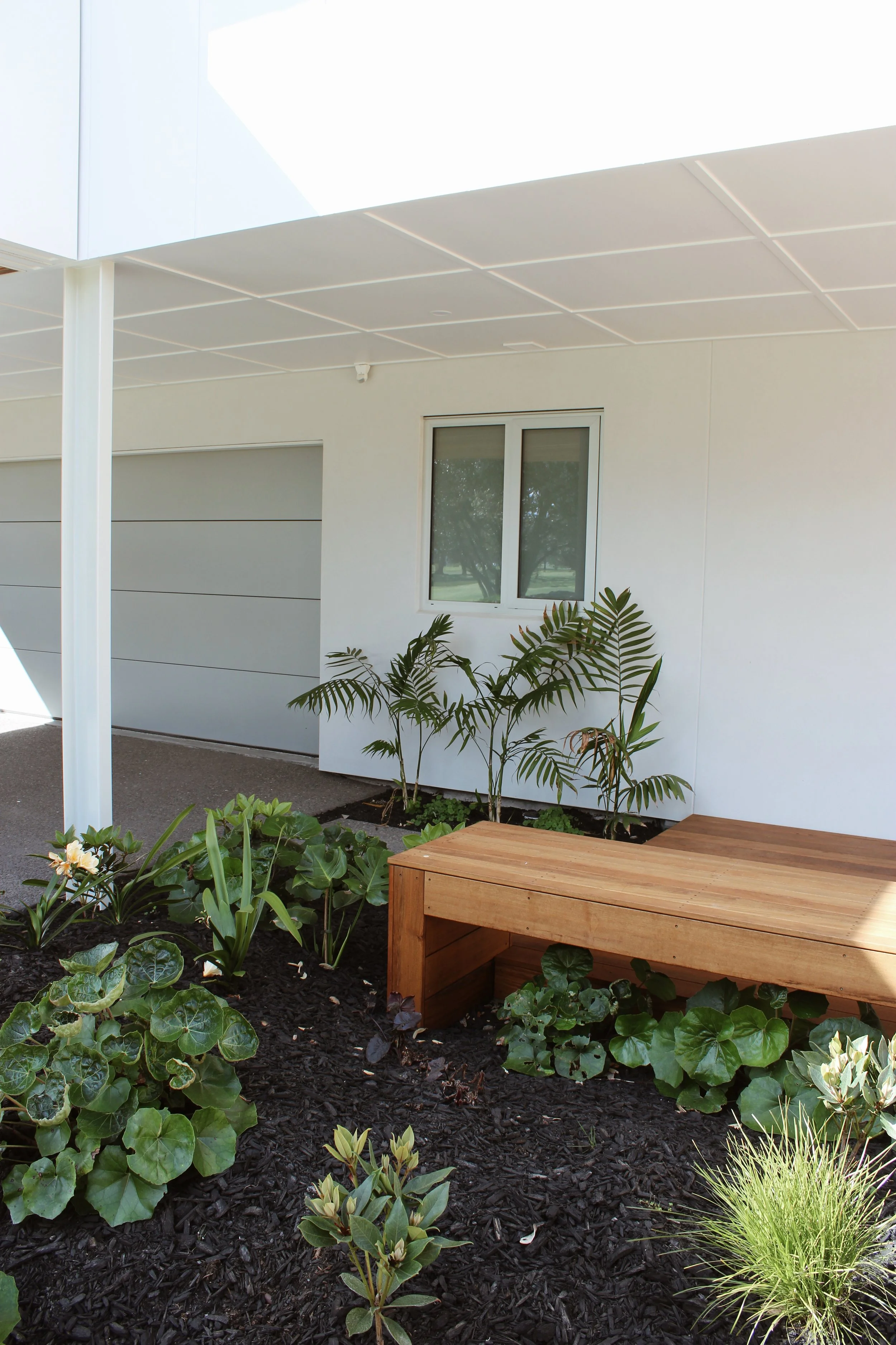 View of a modern house exterior with white walls, a window, indoor plants, and a wooden outdoor bench, with a garden bed filled with various green plants and mulch.