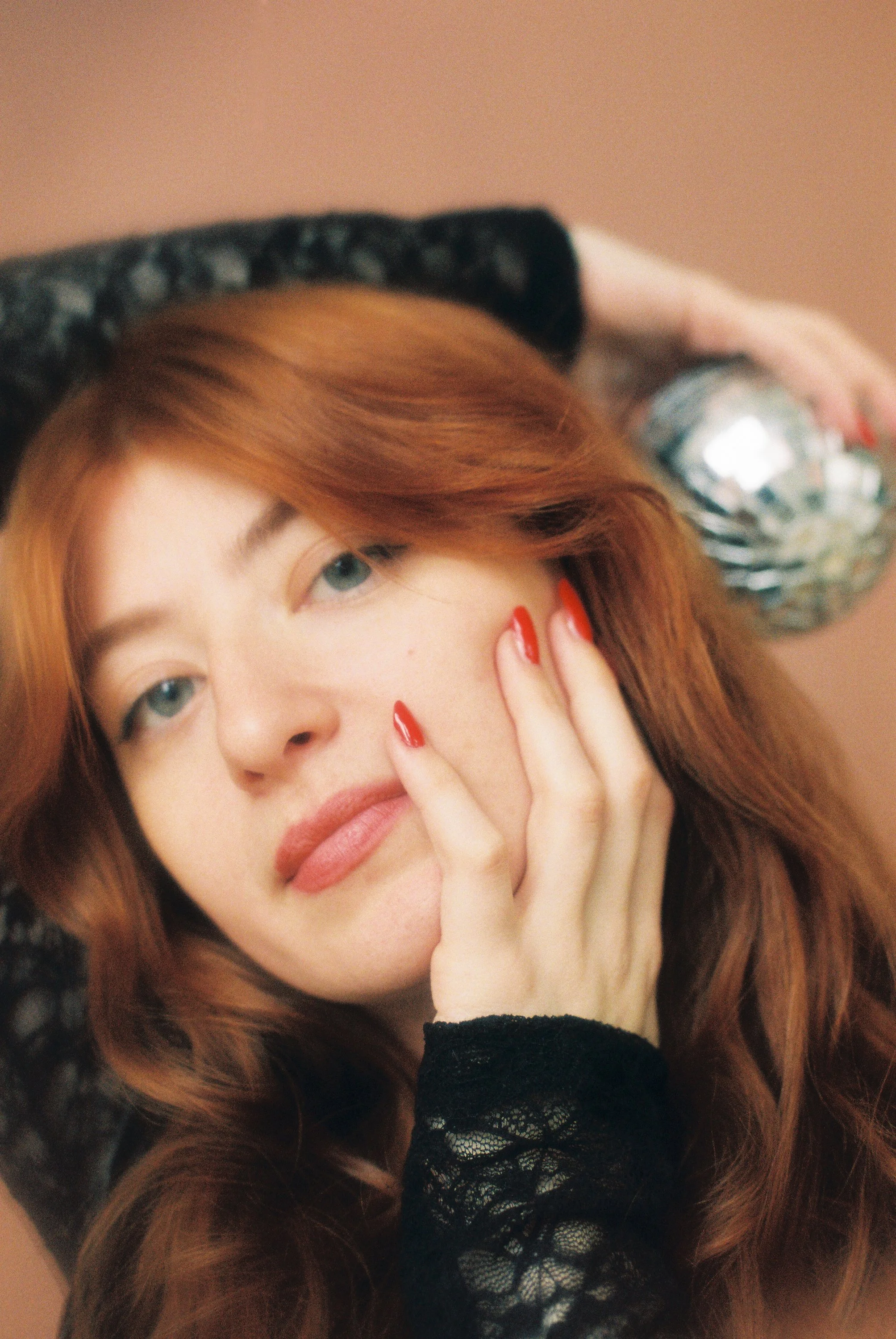 A person with long, wavy red hair partially covering their face, wearing black lace clothing, and holding a shiny, reflective object near their temple, posing against a muted background.