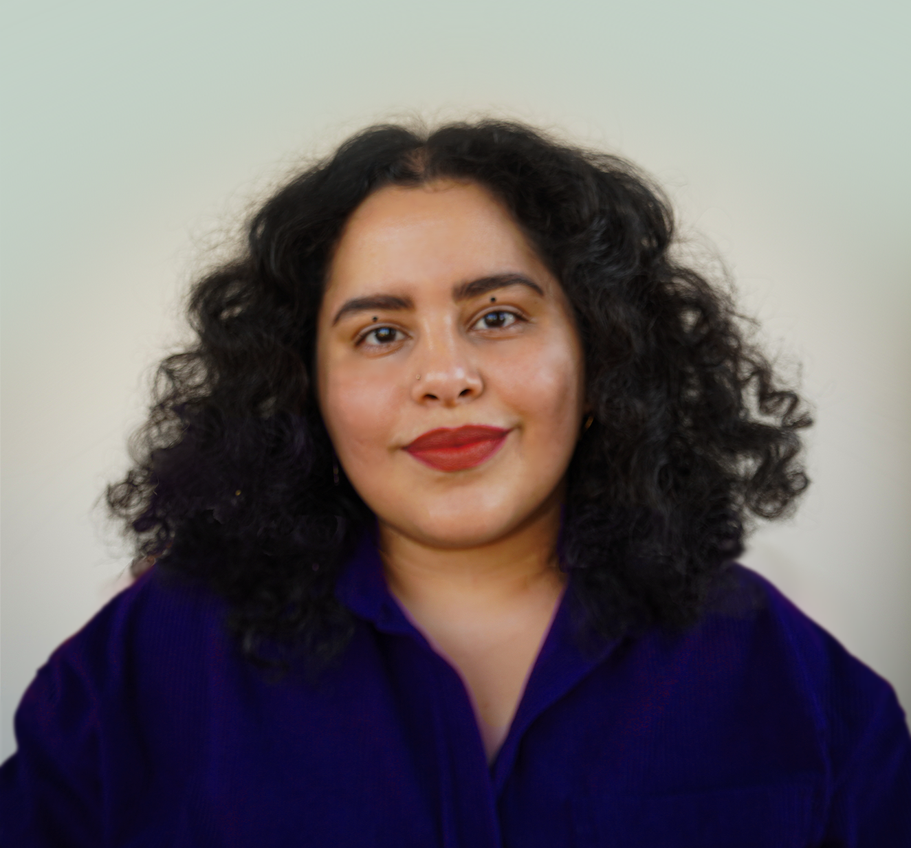 A person with bobbed, wavy black hair parted in the middle, wearing a blue button down top, red lipstick and two dots on her eye lids.