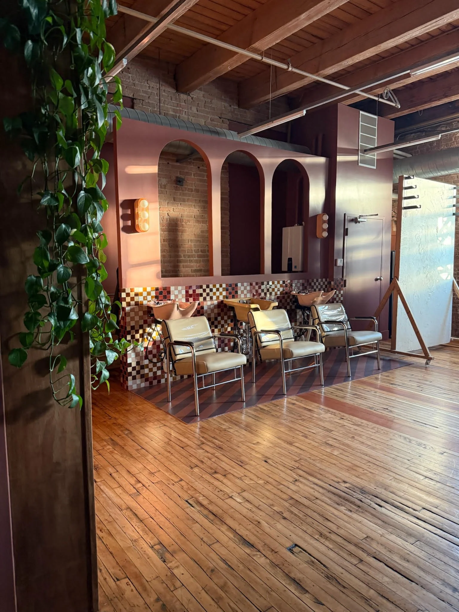 Interior of a modern salon or spa with a wooden floor, exposed brick wall, and a decorative brown and white tiled washbasin area. There are four chairs with armrests in front of the washbasin and a bunch of green leafy plants on the left side.
