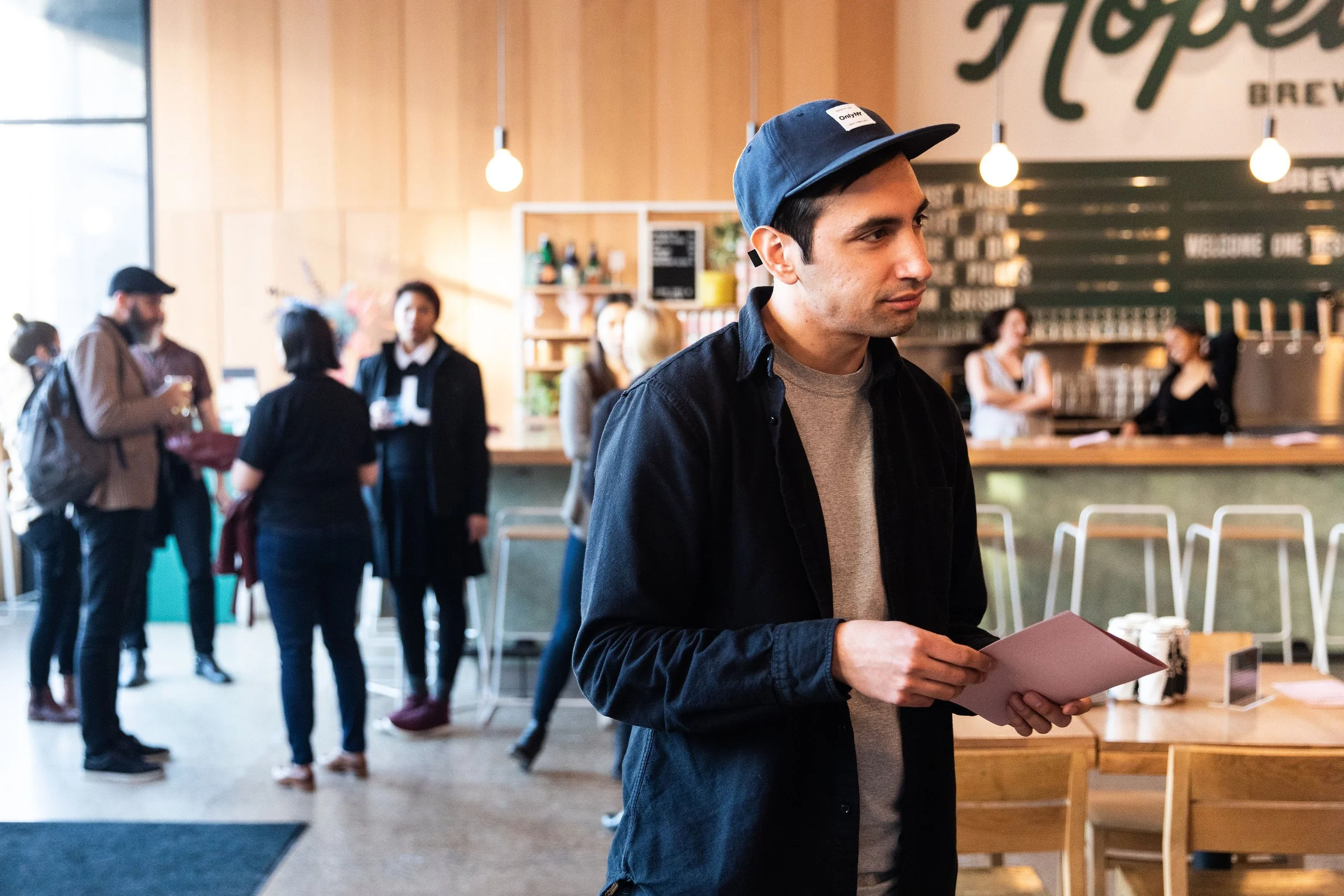 A man in a blue cap and black jacket holding a menu or flyer in a coffee shop with several people in line and working behind the counter.