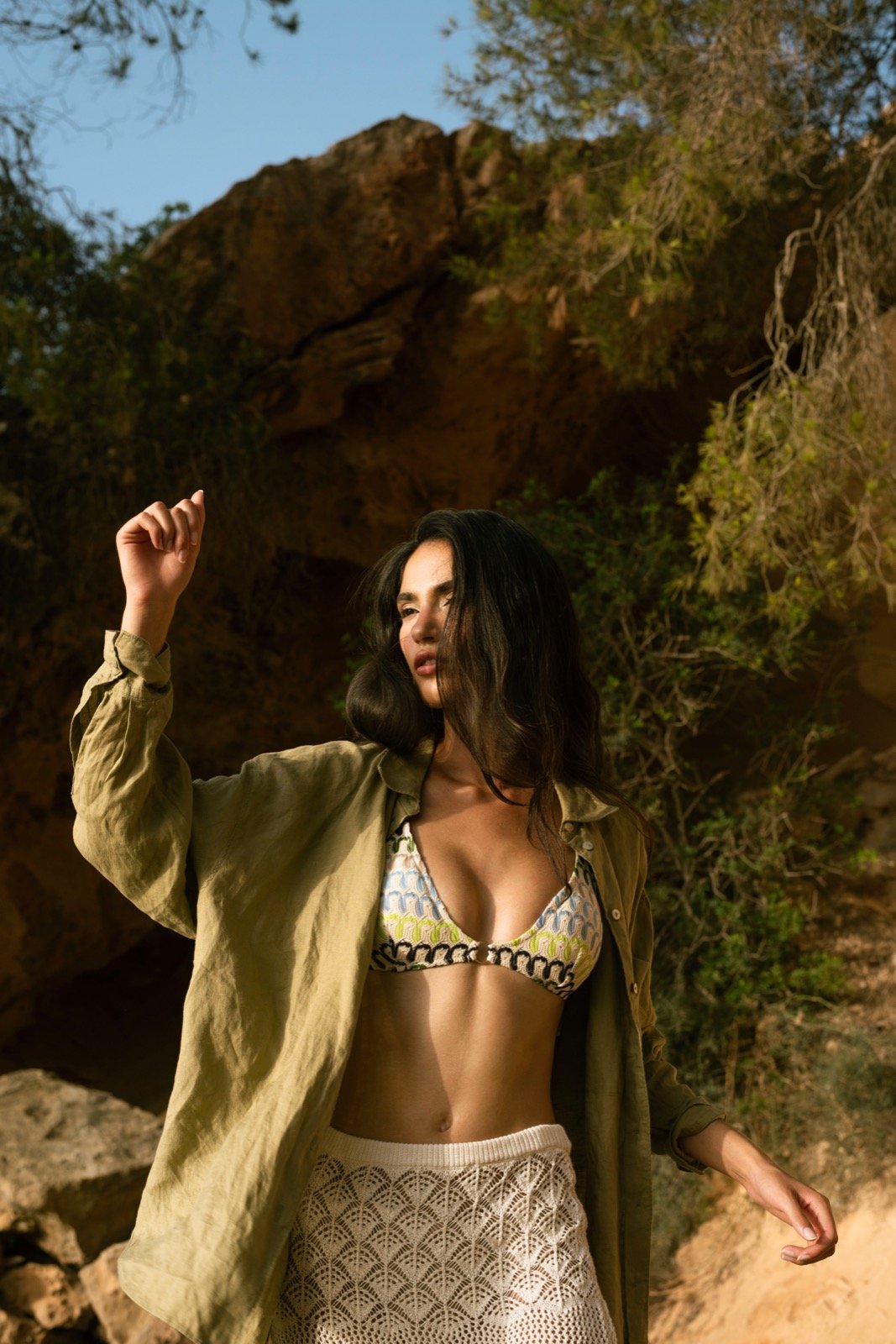 A woman with dark wavy hair wearing a colorful bikini top, light-colored skirt, and an open olive green shirt, standing outdoors near rocks and trees - Model influencer photo-shooting
