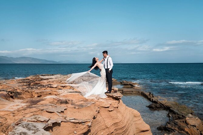 A couple in wedding attire stands on a rocky coast by the ocean, holding hands and looking at each other.