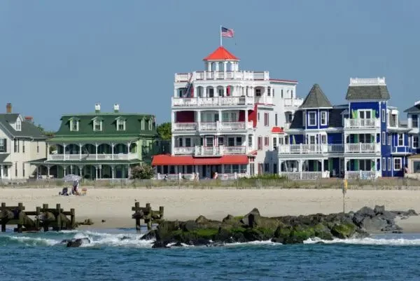 Colorful beach houses along the shoreline, including a large white house with red accents and an American flag on top, with a sandy beach and rocks in the foreground.