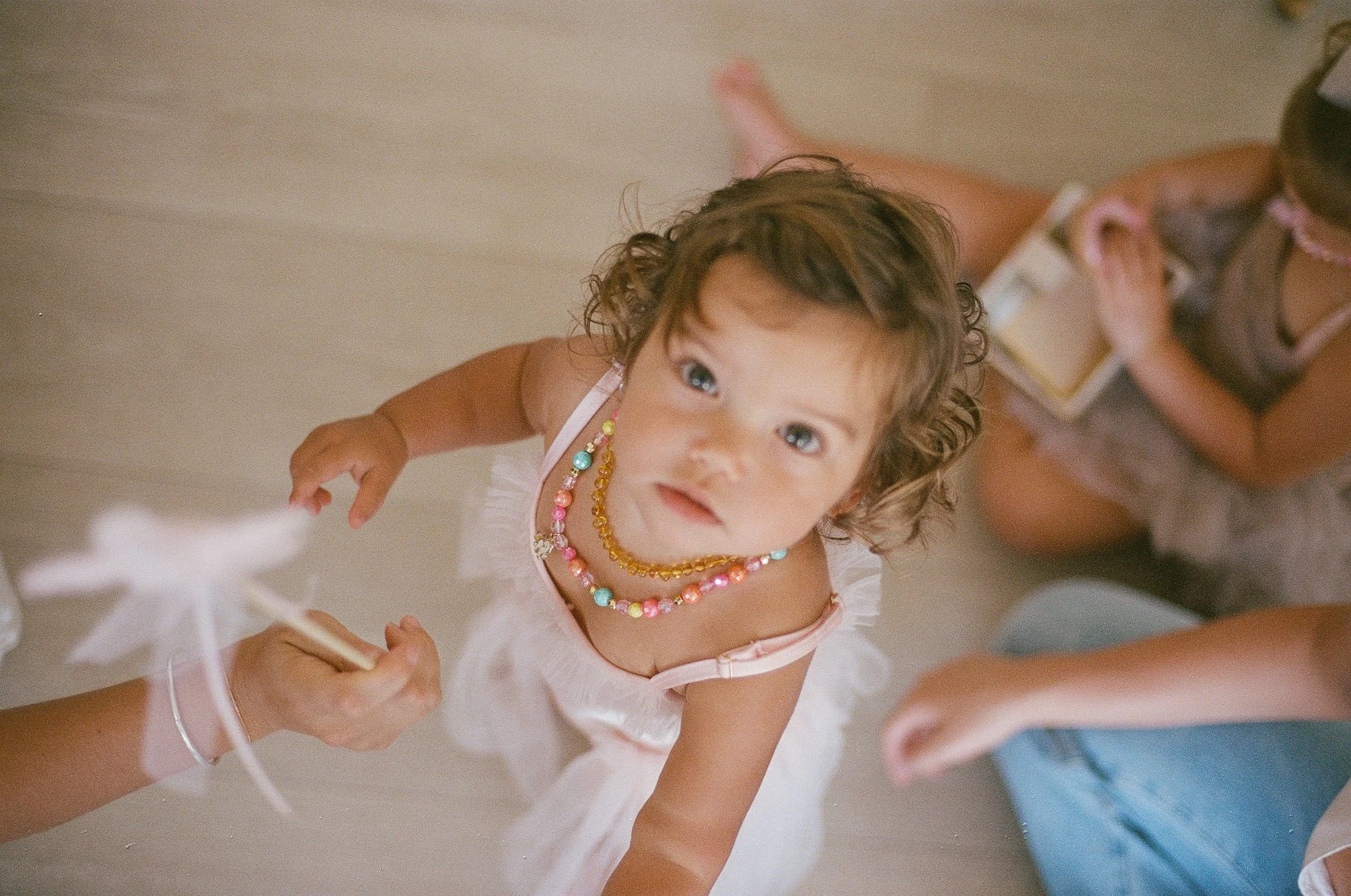 Young girl with curly hair and blue eyes, wearing colorful beaded necklaces and a light pink dress, looking up at the camera with a curious expression. She is sitting on the floor with other children partially visible around her.