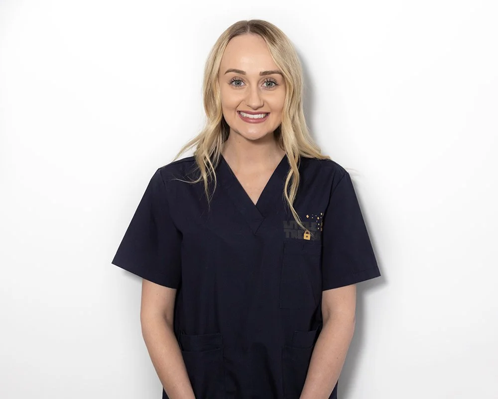 A woman with blonde hair wearing dark medical scrubs, smiling, standing against a plain white background.