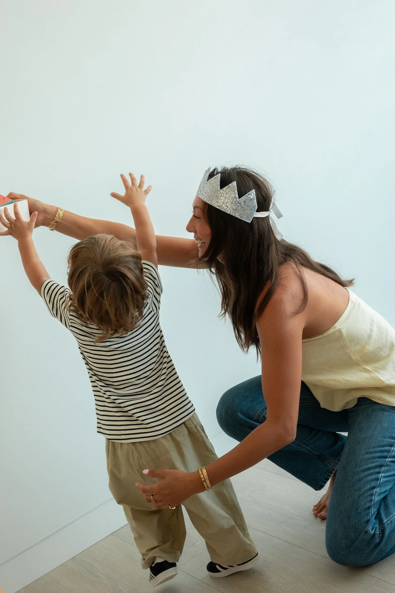 A woman wearing a silver party crown is playing with a young child indoors against a plain white wall.