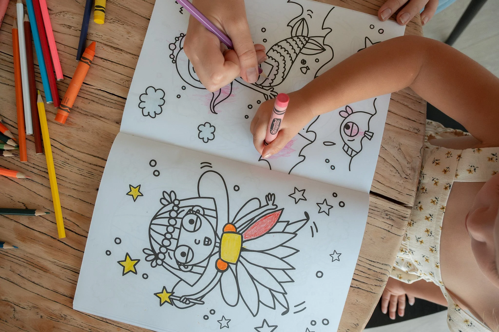 Children coloring a fairy-themed coloring book with crayons and pencils on a wooden table.