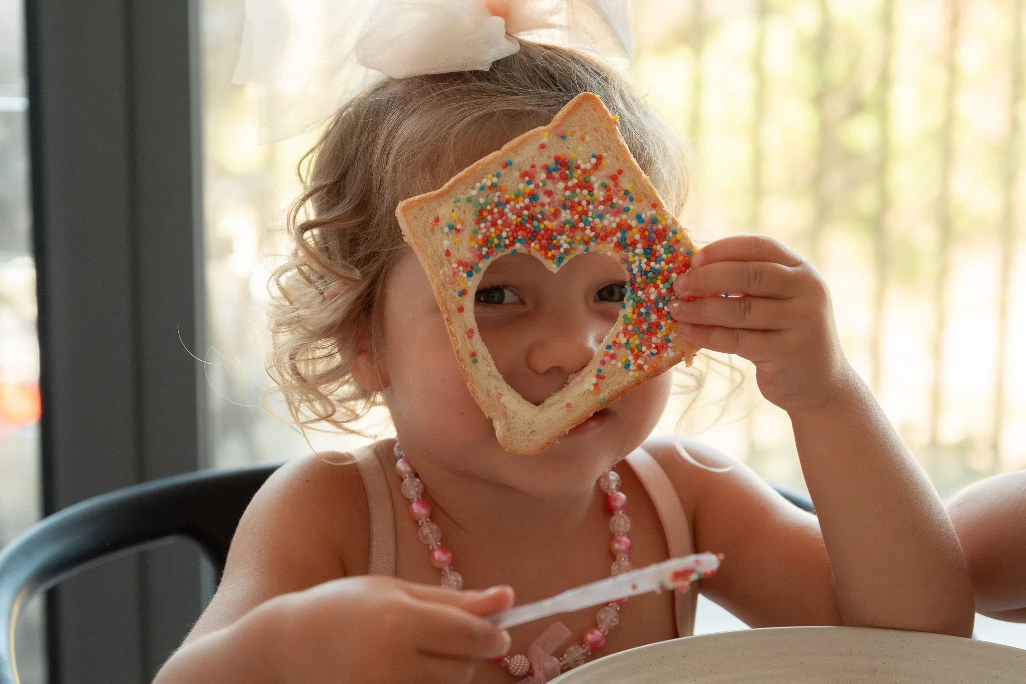 A young girl with curly blonde hair wearing a pink necklace and pink dress holding a slice of bread shaped like a heart with colorful sprinkles, covering her right eye, inside a room with sunlight and trees outside the window.