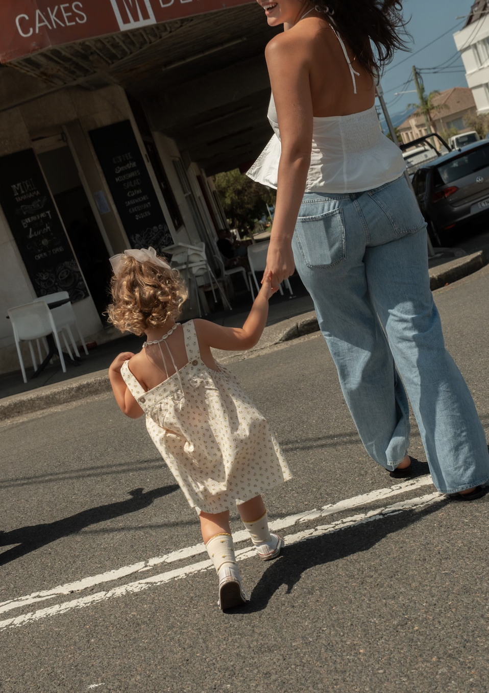 Nanny holding hands with a little girl in a dress walking on a street near a café.