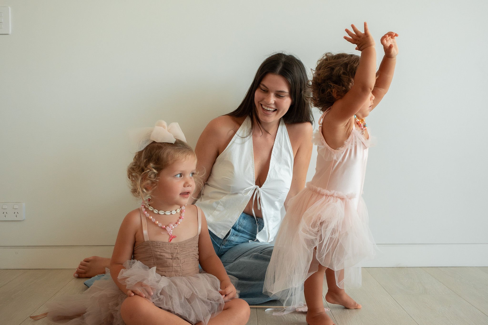 Woman and two young children in ballet dresses sitting on a wooden floor, smiling and playing.