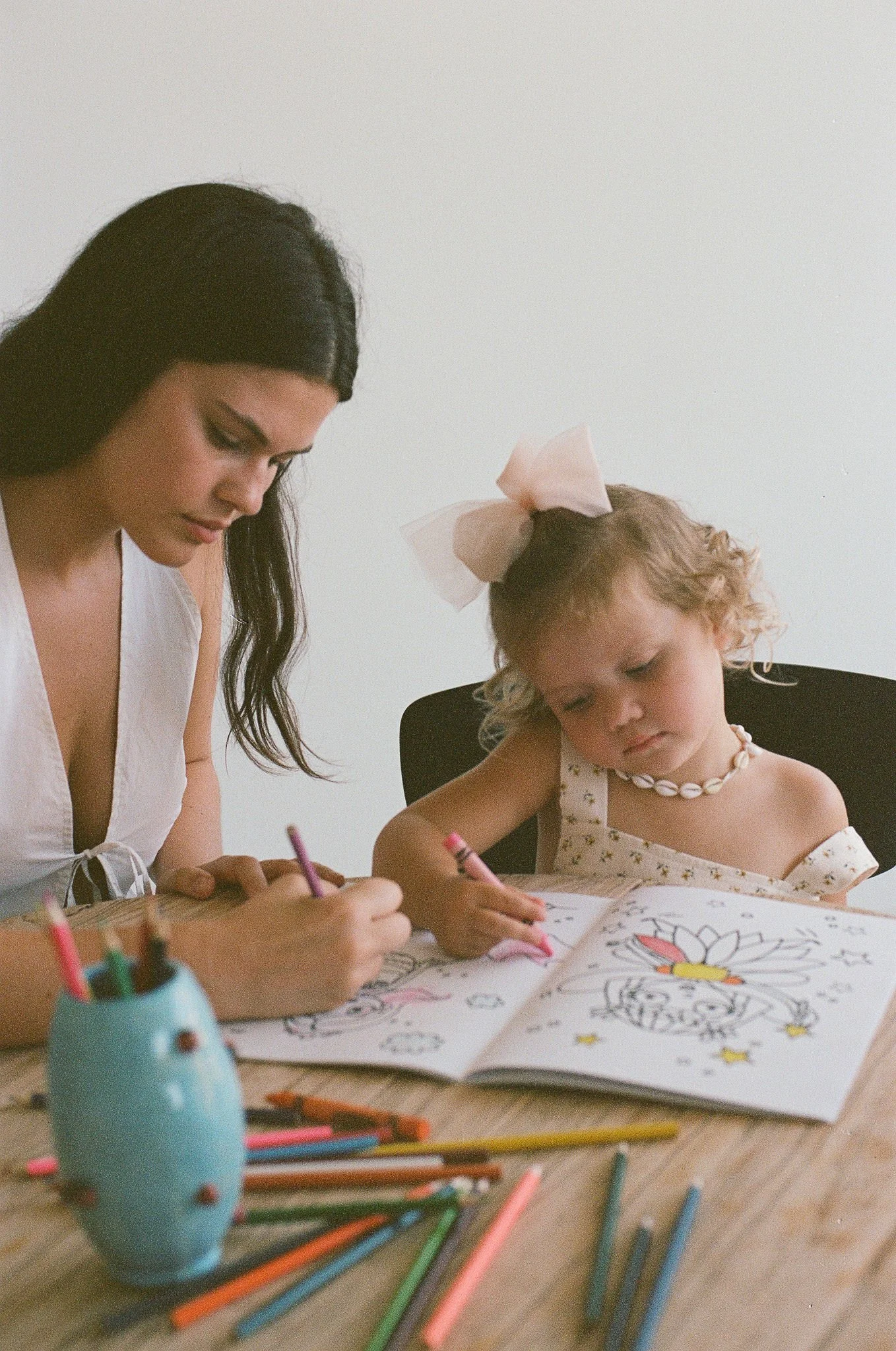 A woman and a girl drawing coloring pages at a wooden table. The girl has a large pink bow in her hair and wears a shell necklace. The table has colored pencils and a blue pencil holder.