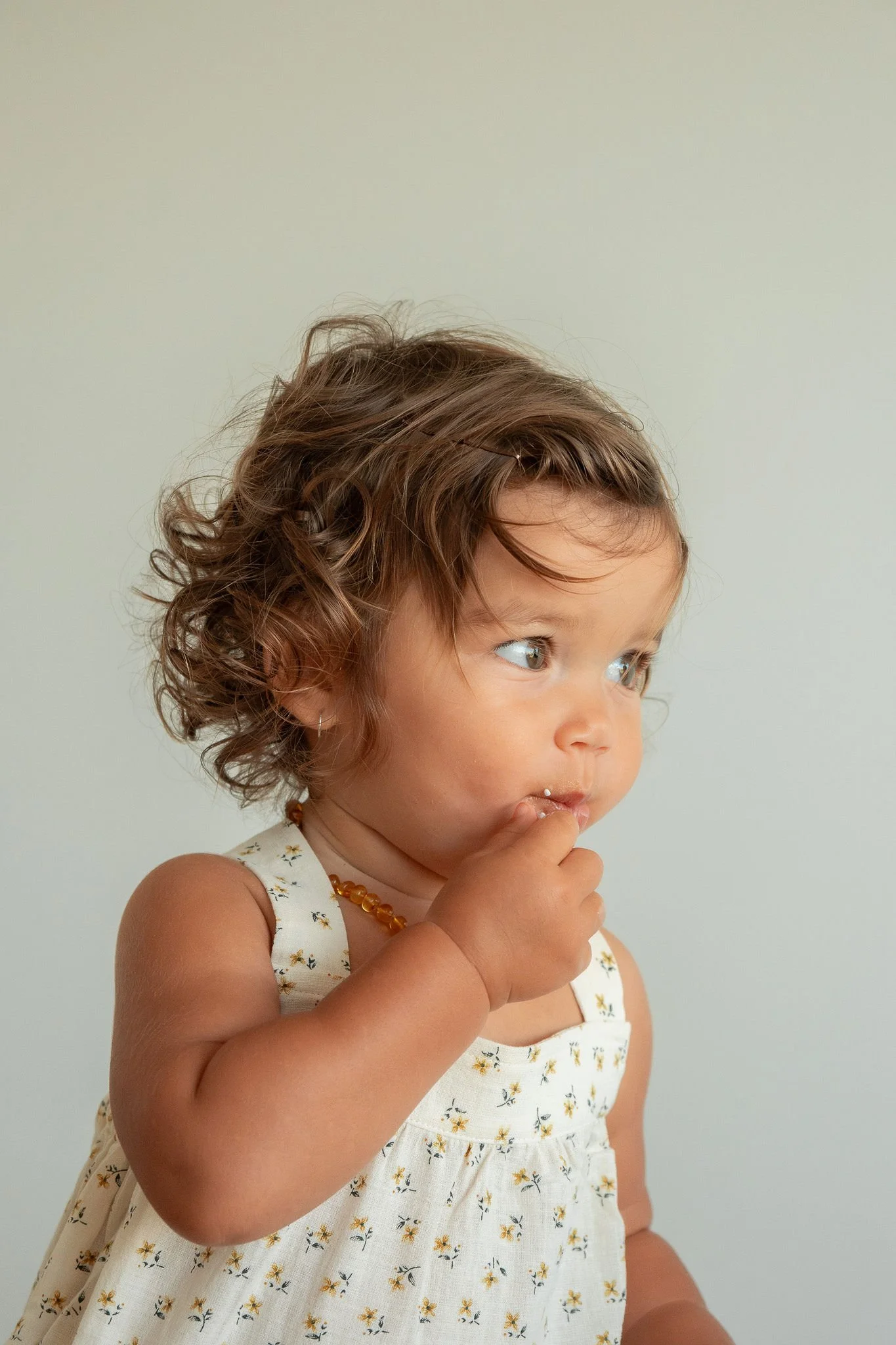 Young girl with curly brown hair, wearing a white dress with small yellow flowers, and a beaded necklace, is touching her lips and looking to her right against a plain light background.