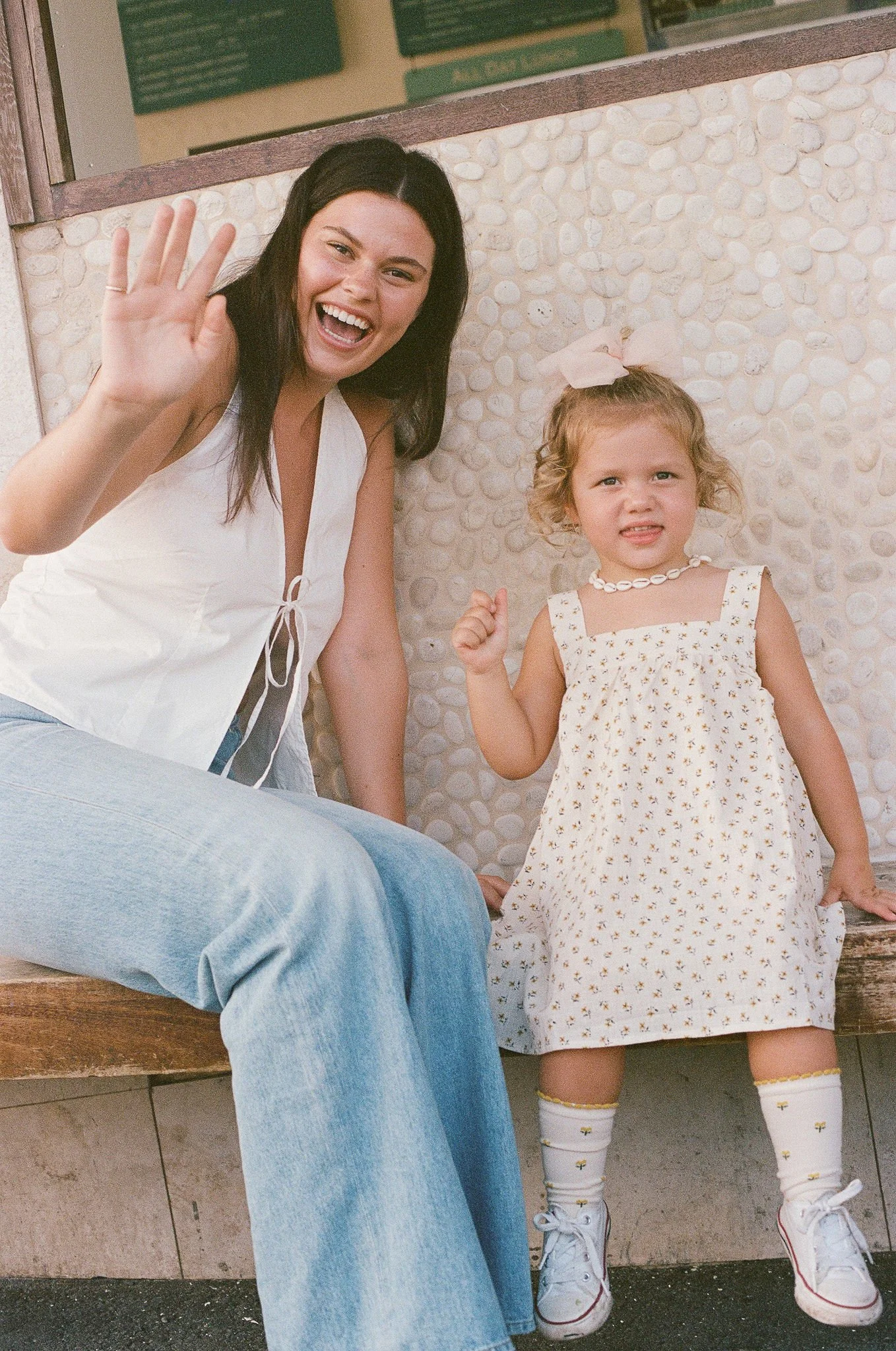 Nanny and litte girl smiling and sitting on a bench, with the woman waving at the camera. The girl is wearing a white dress and sneakers, while the woman is dressed in a white sleeveless top and jeans.