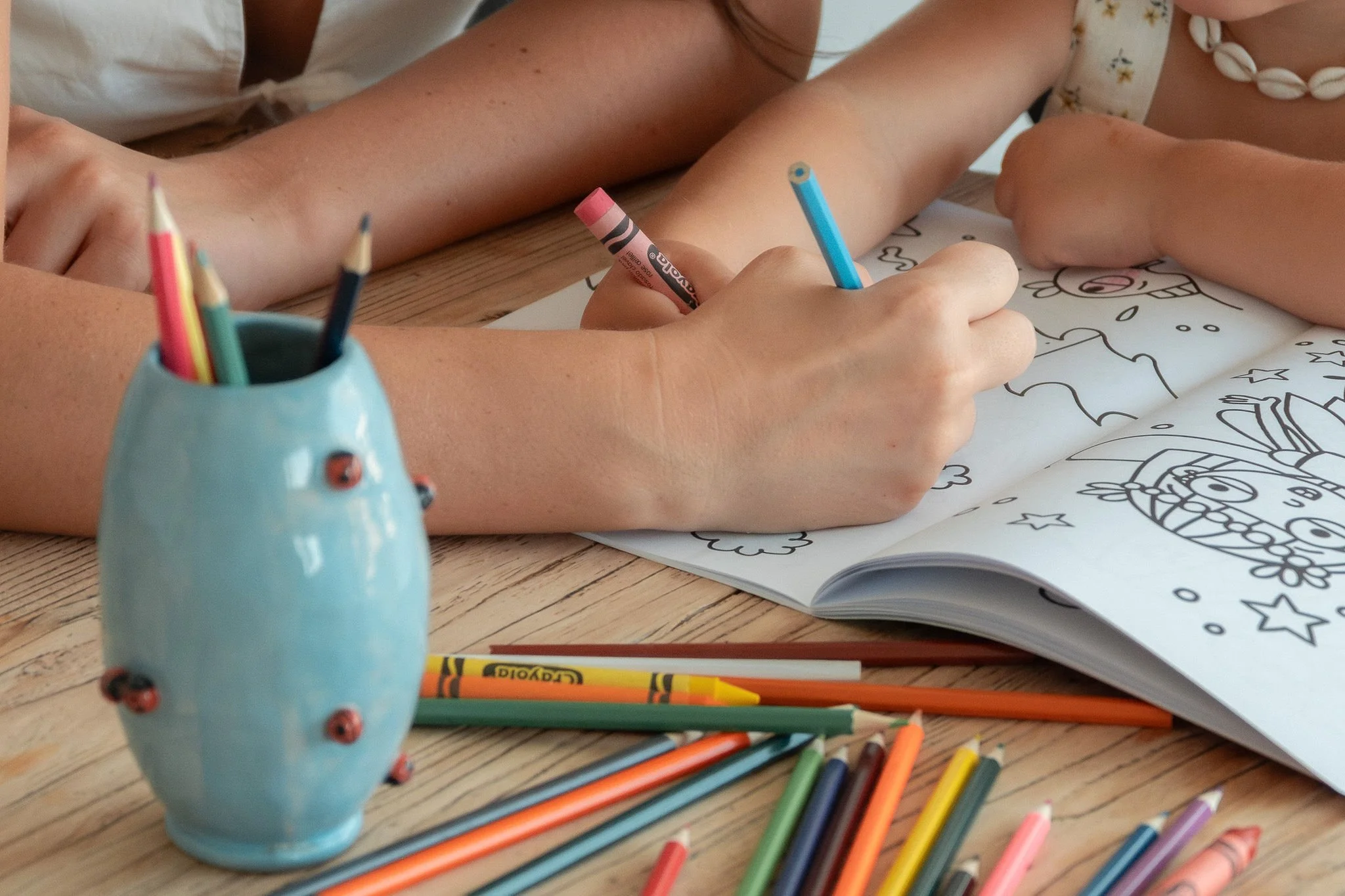 Two children drawing and coloring in a coloring book with various colored pencils and crayons on a wooden table.