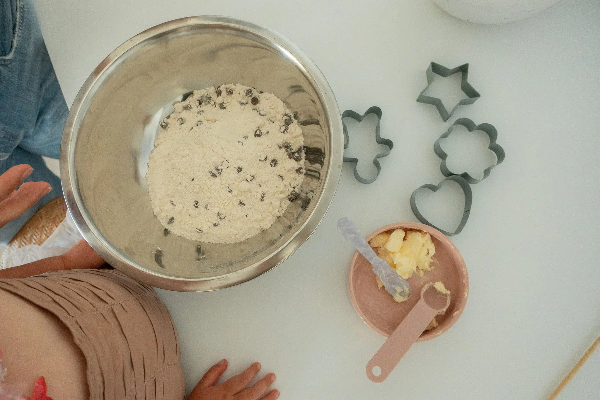 A child with a blue scooter and an adult with a bicycle, standing on a brick path surrounded by greenery in the background.