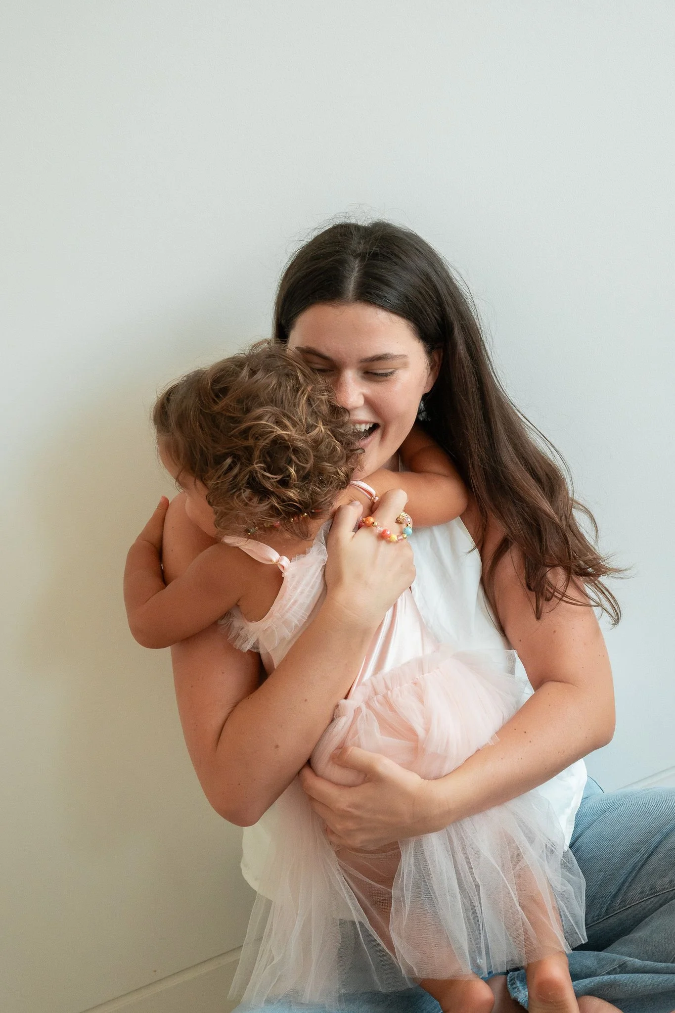 A woman holding a young girl, both smiling and hugging each other affectionately indoors.