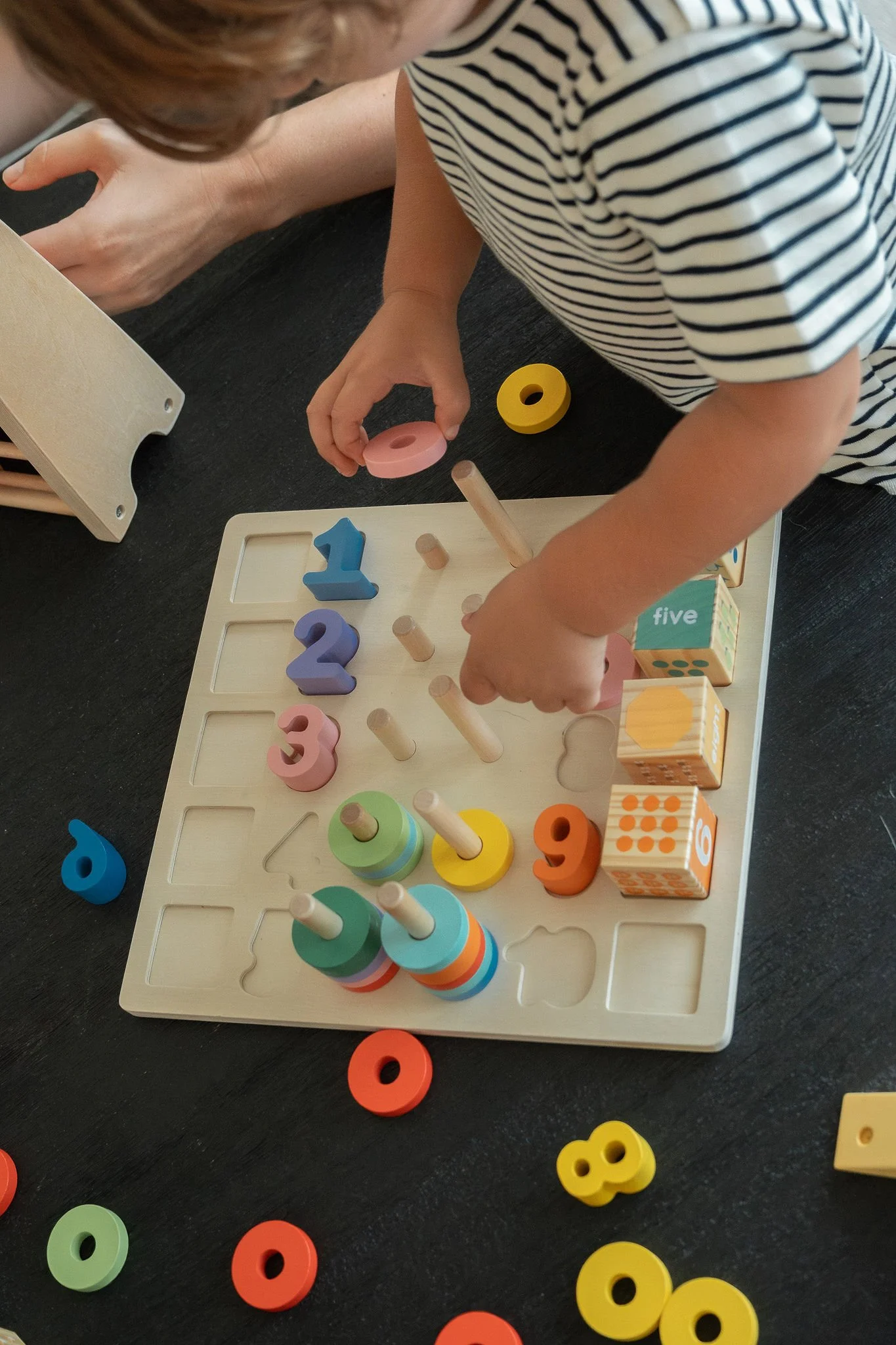 A child playing with a wooden educational toy that has numbered and colored pieces, some stacking and some fitting into the puzzle board, on a black surface.
