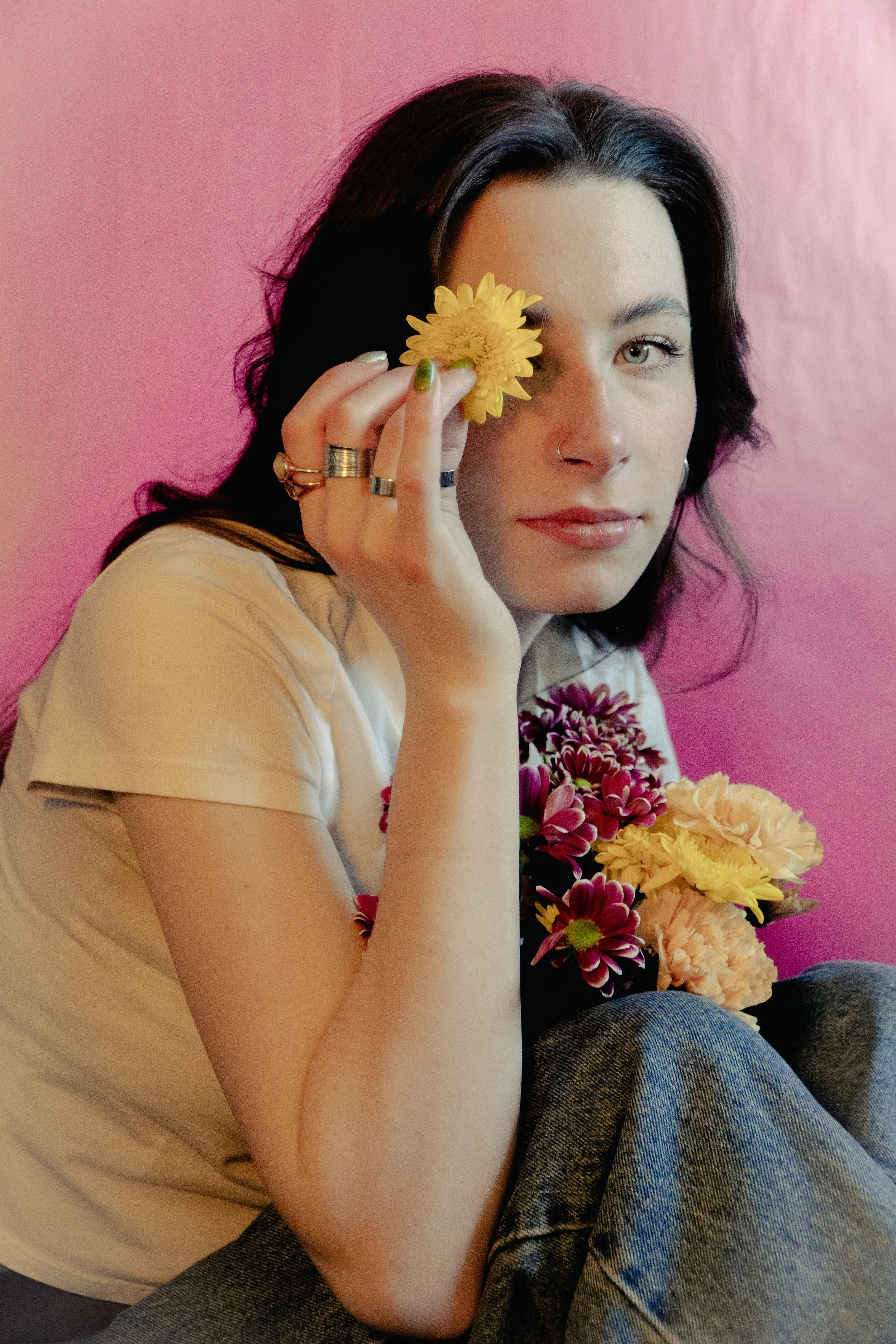 Woman holding flowers with pink background, wearing a white shirt and jeans, holding a yellow flower near her eye.