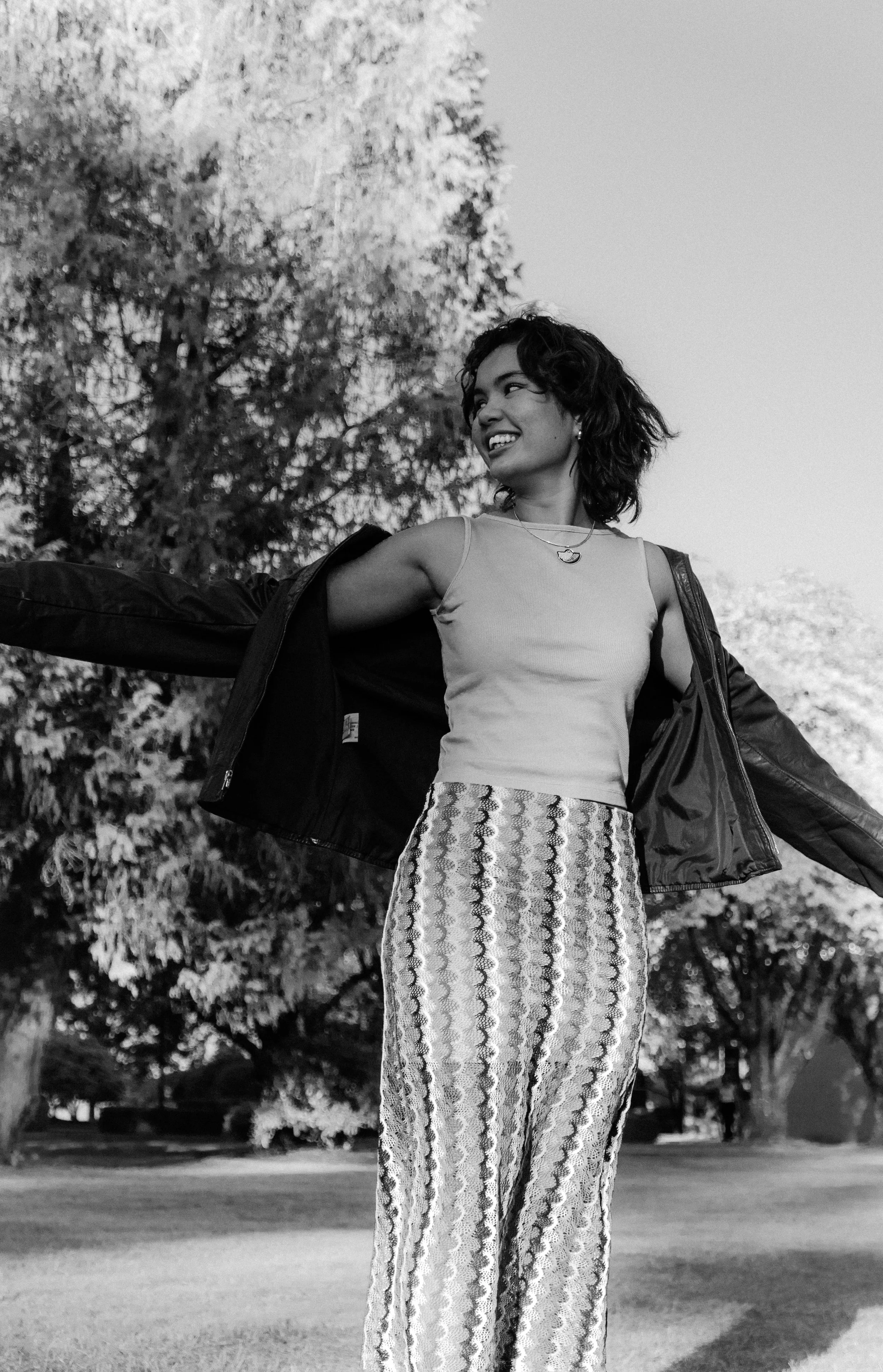 Smiling person outdoors, wearing a sleeveless top and patterned pants, holding a jacket open, with trees in the background.