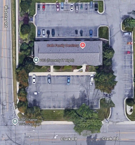 Aerial view of a building and parking lot with designated areas for cars. The building is labeled Bath Family Dentistry and VC3 (formerly IT Right). The parking lot is partly filled with vehicles and is bordered by trees and green spaces. There is a township hall nearby, and the area is bordered by Clark Road and Webster Road.