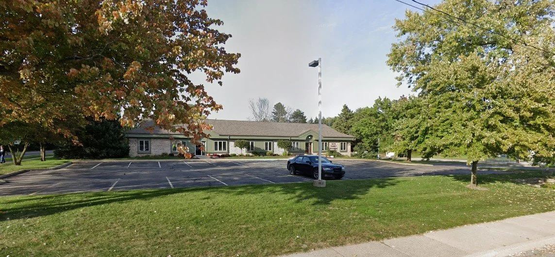 Front view of the building. A parking lot in front, a black car parked near the entrance, trees with autumn foliage, and an American flag on a flagpole.