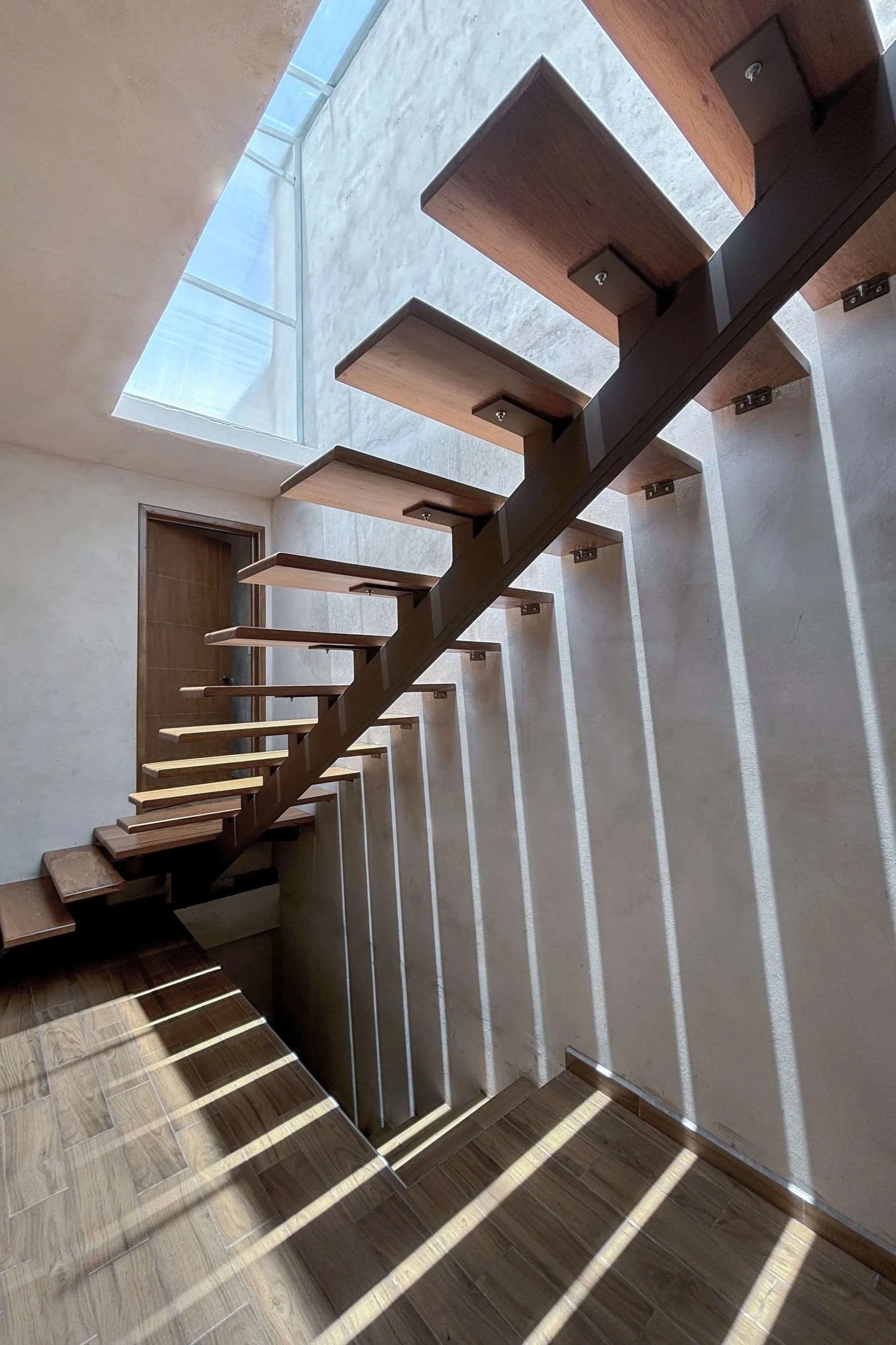 Escalera de madera elegante con estructura metálica negra, con luz natural que crea sombras en la pared y el piso de madera.