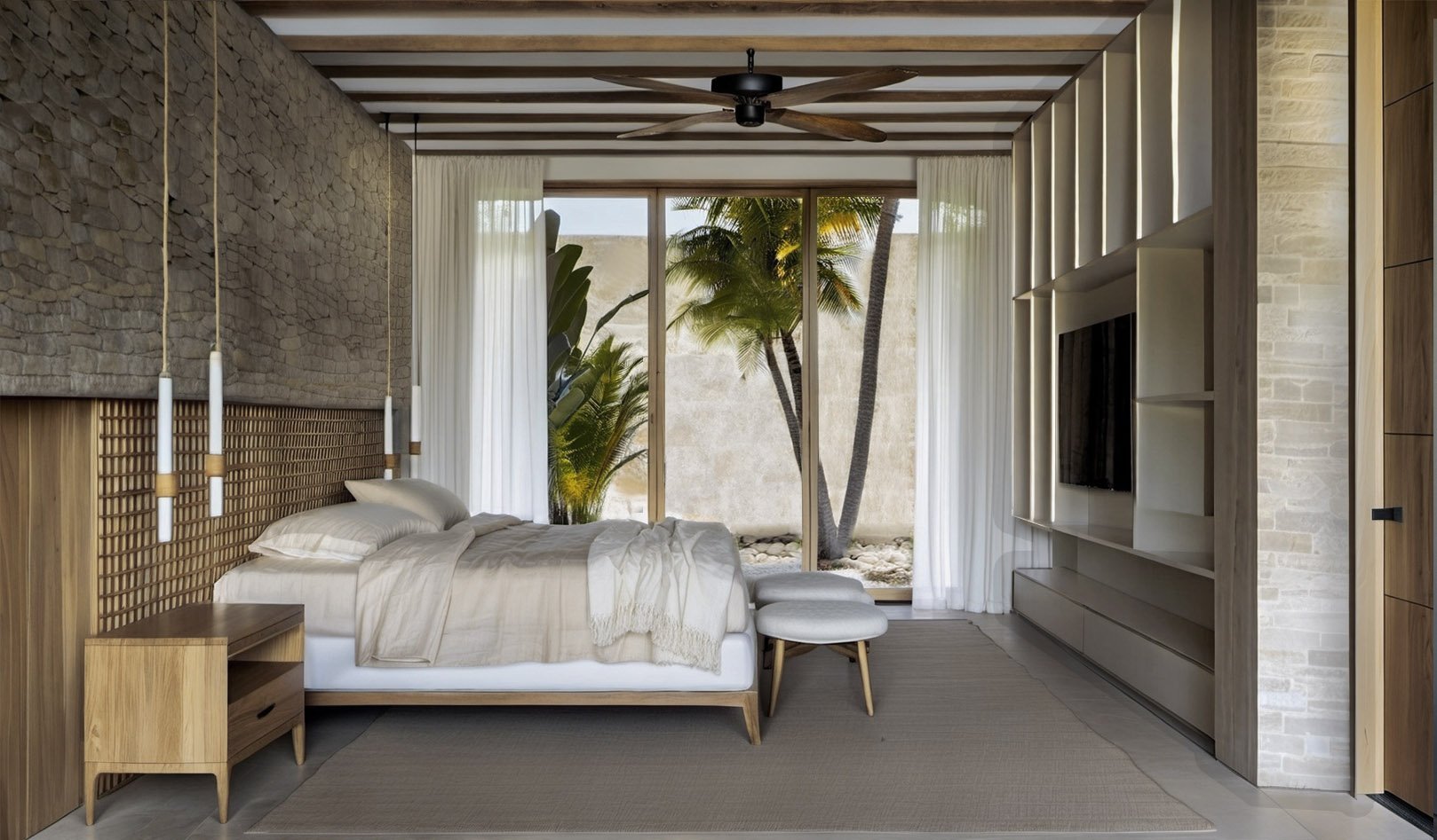 Dormitorio con vista a un jardín con palmeras, paredes de piedra y madera, cama con ropa blanca, cortinas blancas, iluminación suave, ventilador de techo de madera y televisor en una pared de madera