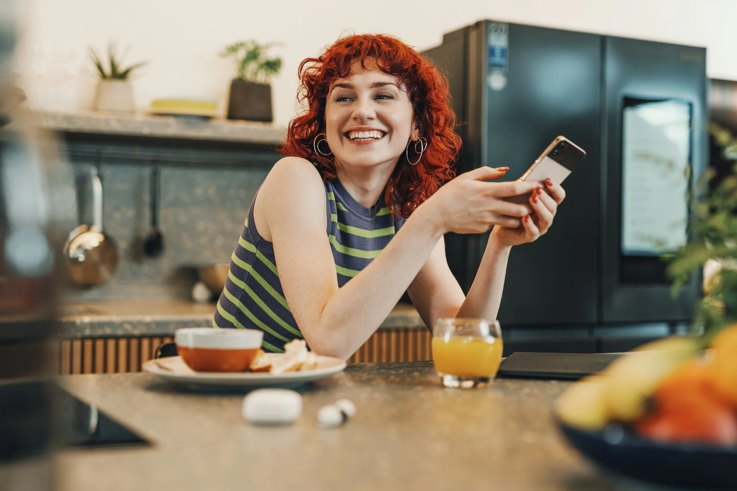 A smiling woman with curly red hair wearing a striped sleeveless top, sitting at a kitchen counter while holding a smartphone.