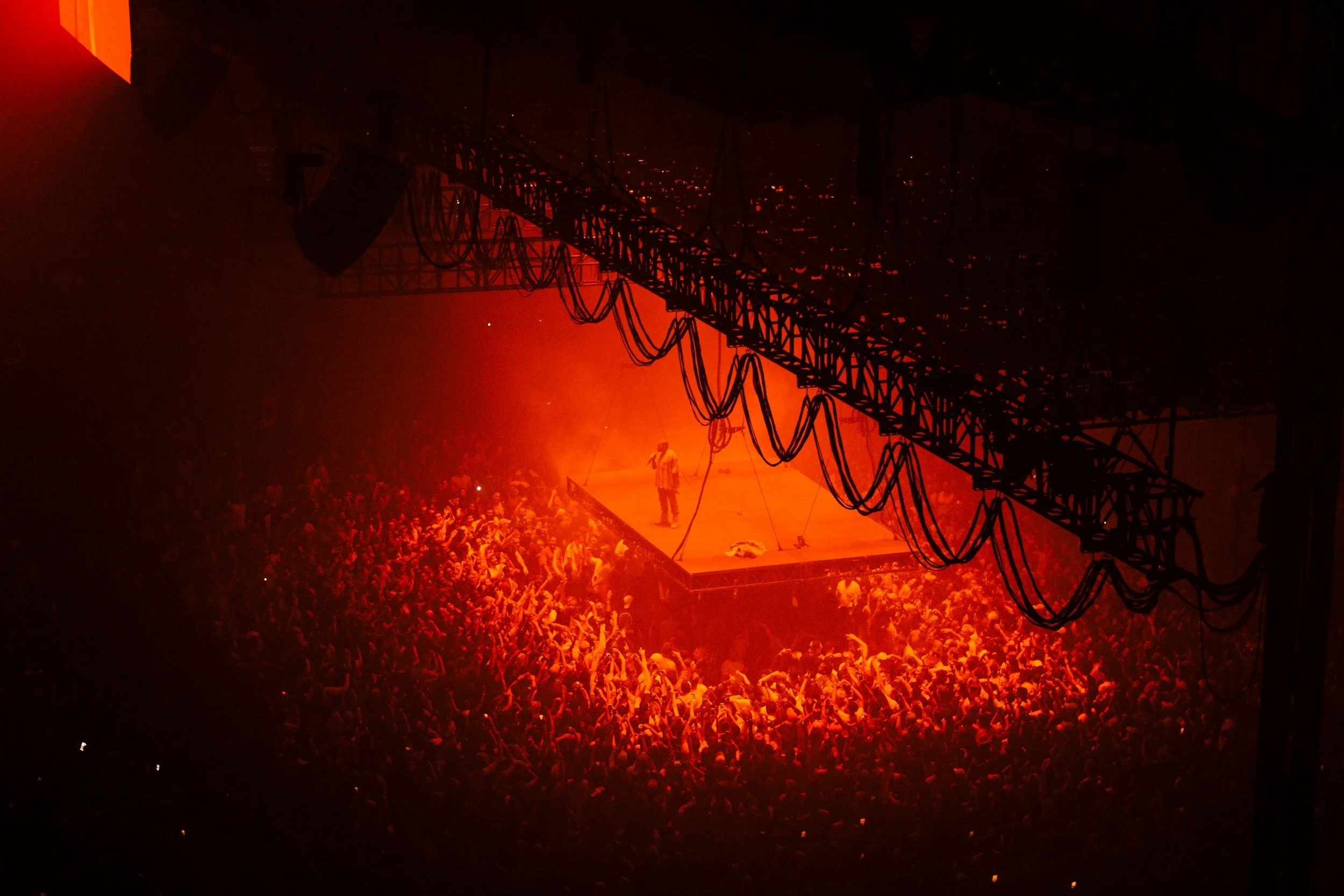 Concert crowd with a performer on stage illuminated in orange light, seen from above with stage rigging overhead.