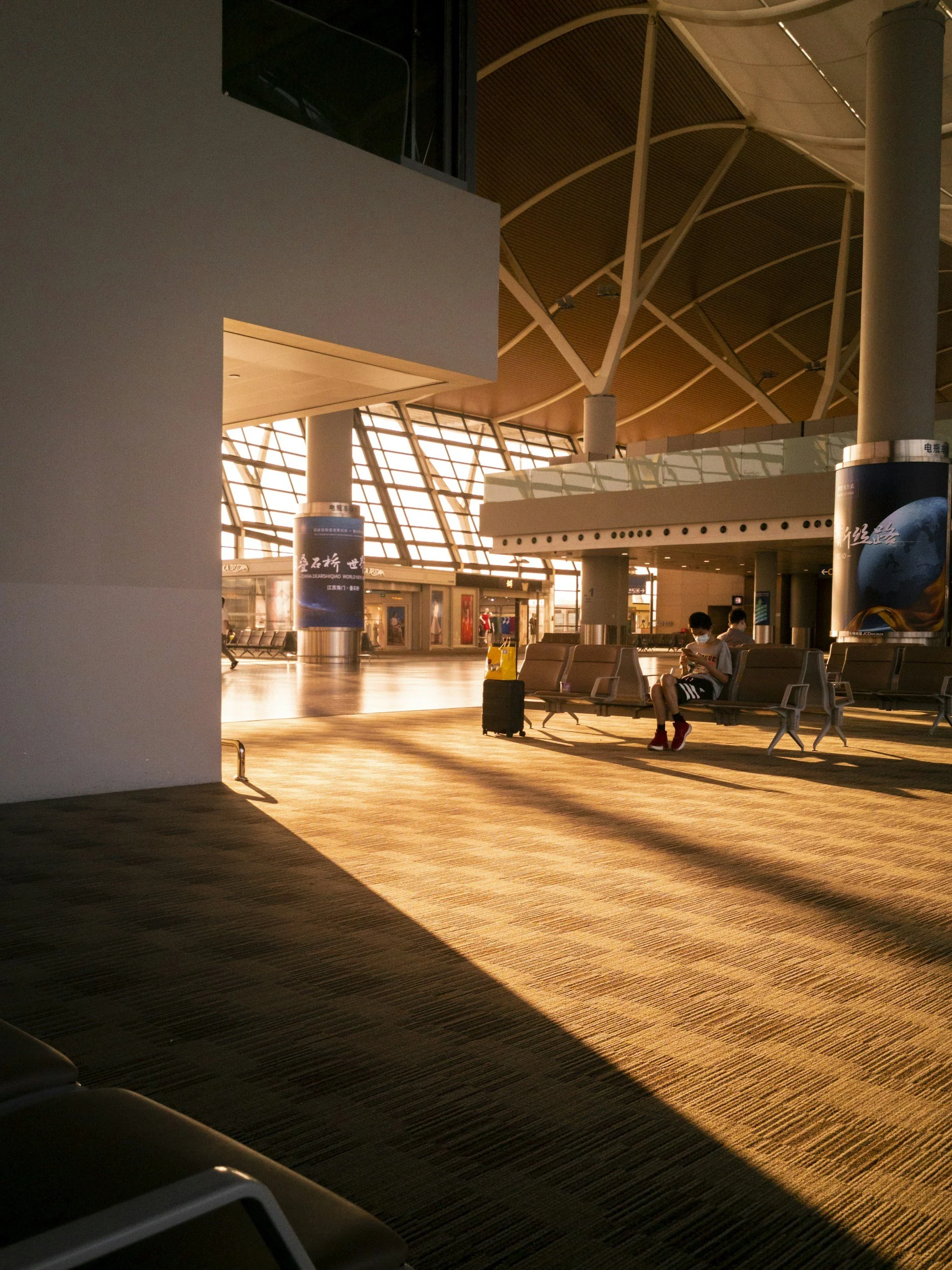 Interior view of an airport terminal with sunlight streaming through large windows, casting long shadows on the wooden floor. There are empty seats and two people sitting and looking at a device. An airport luggage cart with a small suitcase is nearby.