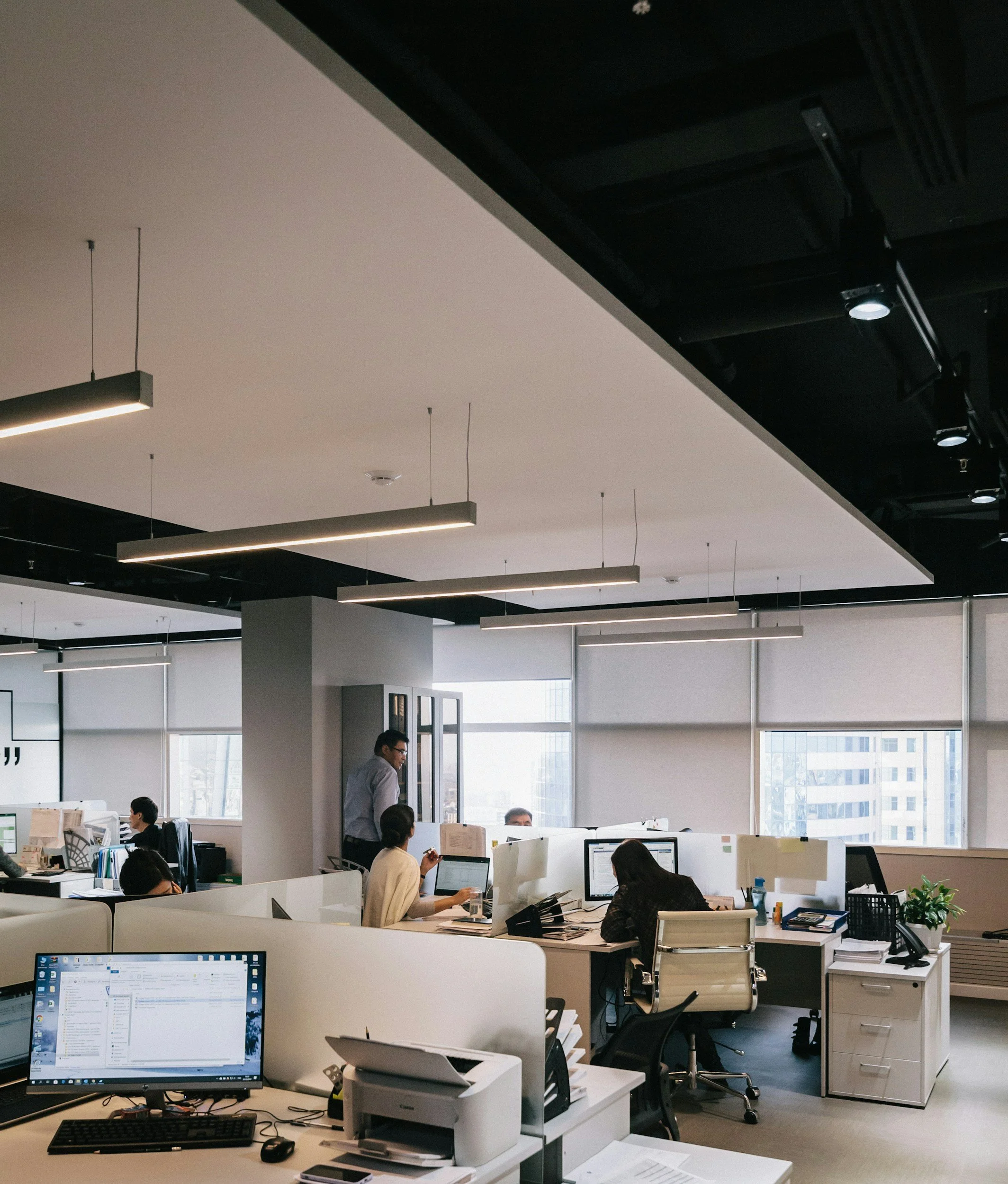 Office with multiple employees working at desks, computers and office supplies, large windows, modern lighting, and high-rise buildings visible outside.