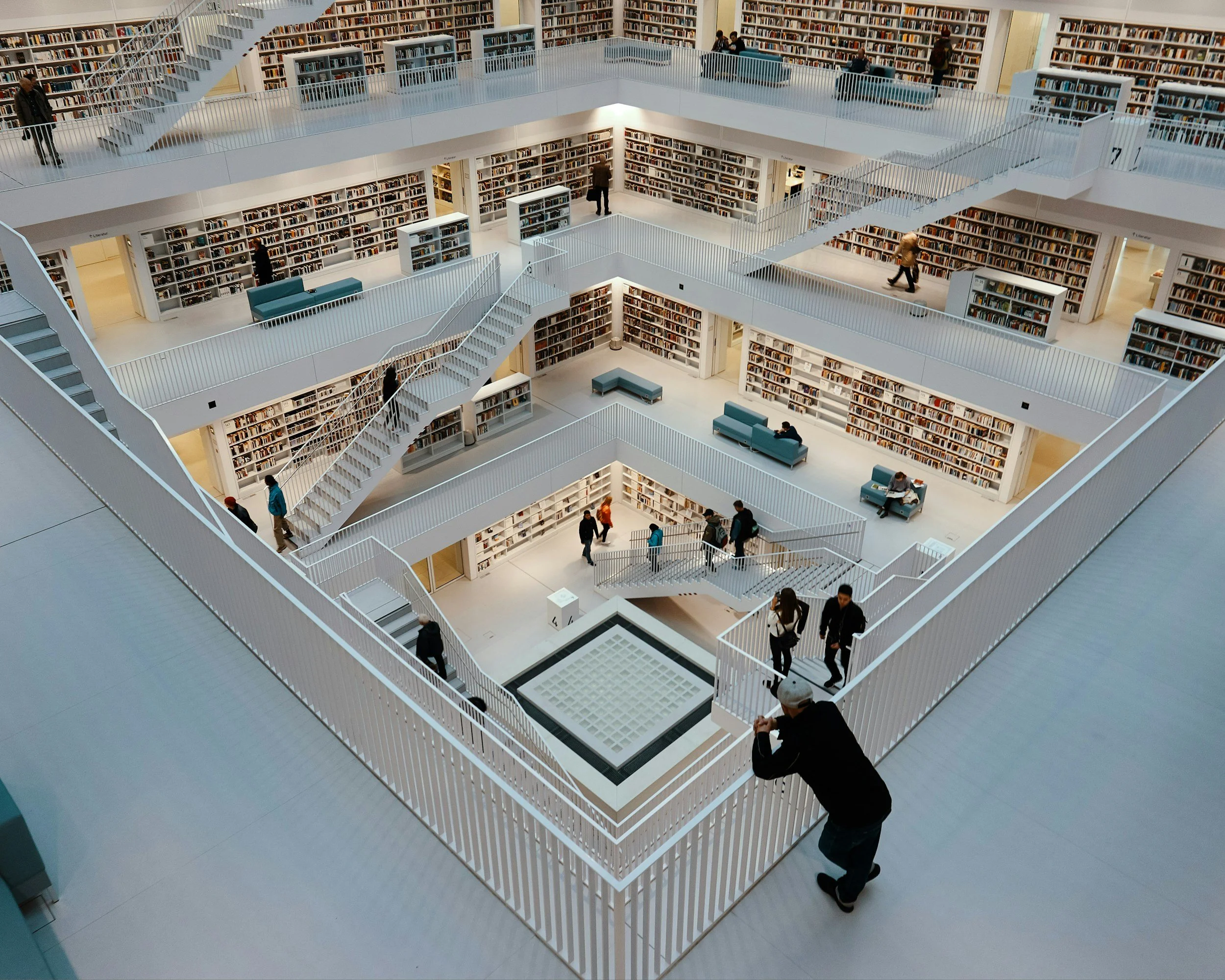 Multiple floors of a modern, multi-level library with white railings, bookshelves filled with books, and people walking and reading.