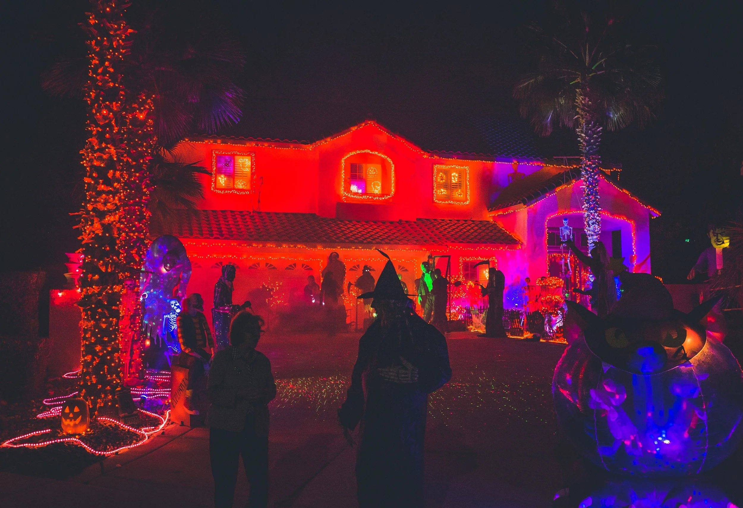 A house decorated with colorful Halloween lights and decorations at night, including pumpkins, skeletons, and witches with people standing in front.