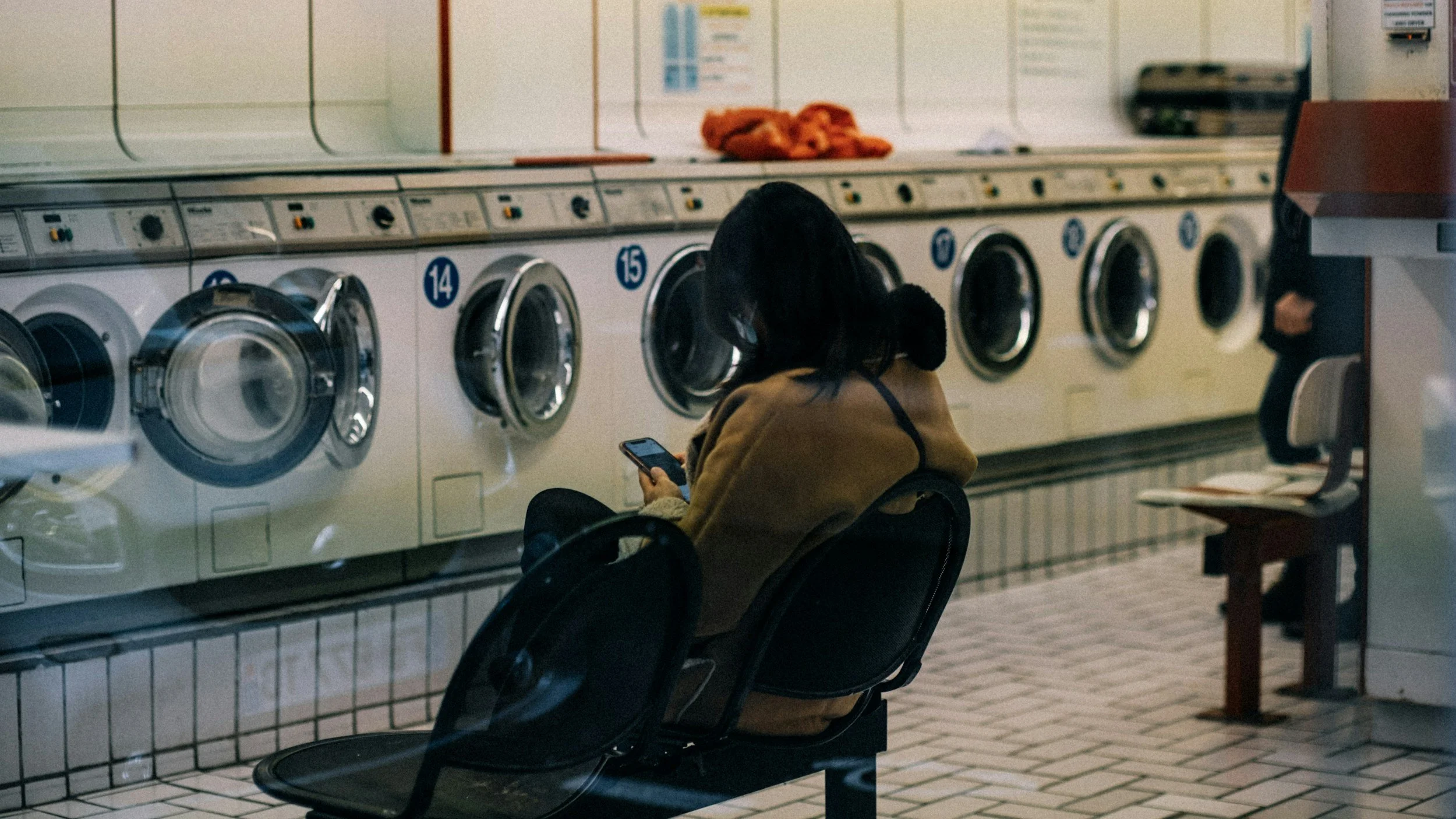 A woman sitting in a laundromat, using her phone while waiting for her laundry to finish. She is seated in front of washing machines lined up against the wall.